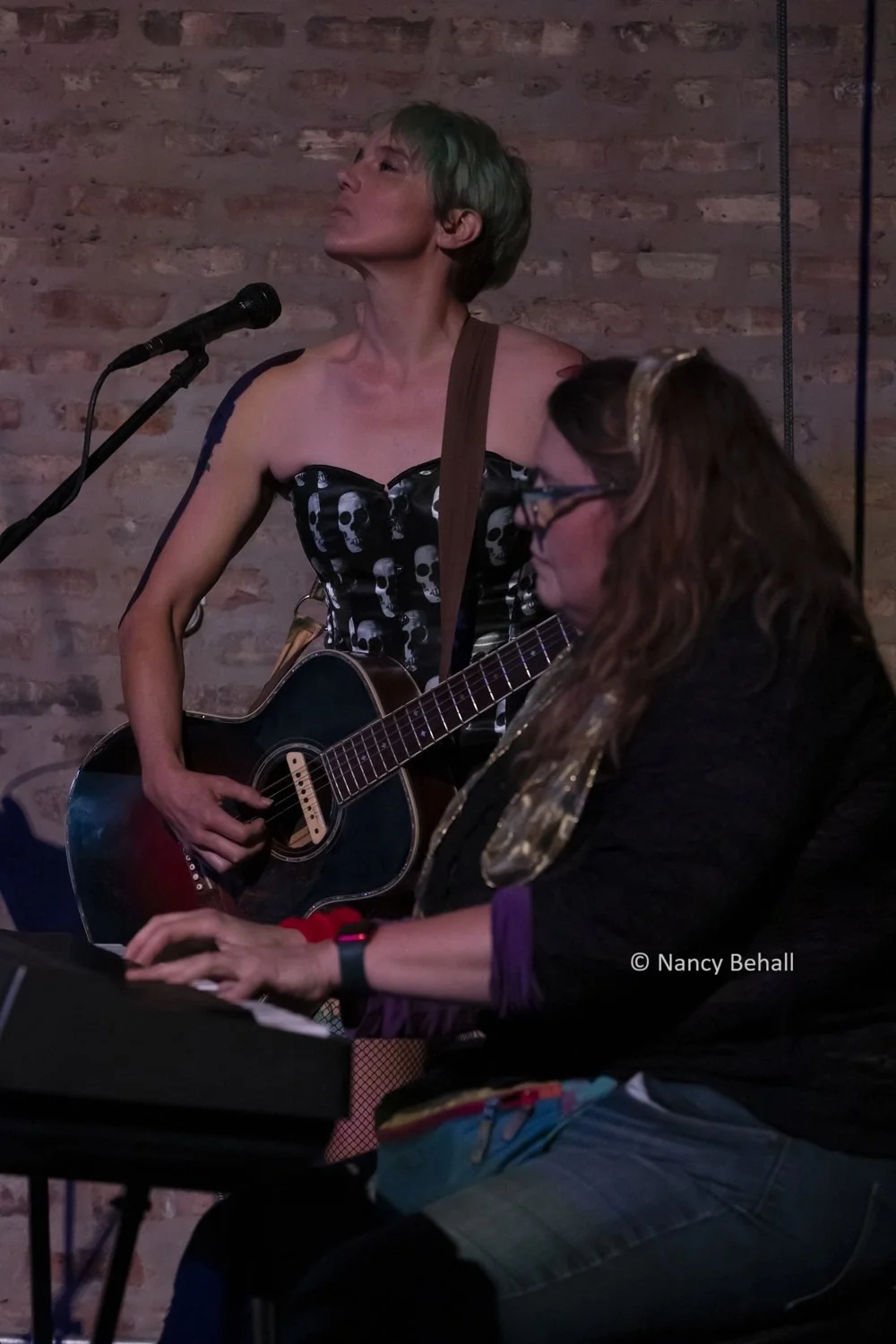 A guitarist and pianist perform on stage against a brick wall. The guitar player wears a sleeveless black skull corset and short green hair and is singing with their eyes closed, while the pianist plays a keyboard, wearing glasses and a dark outfit.