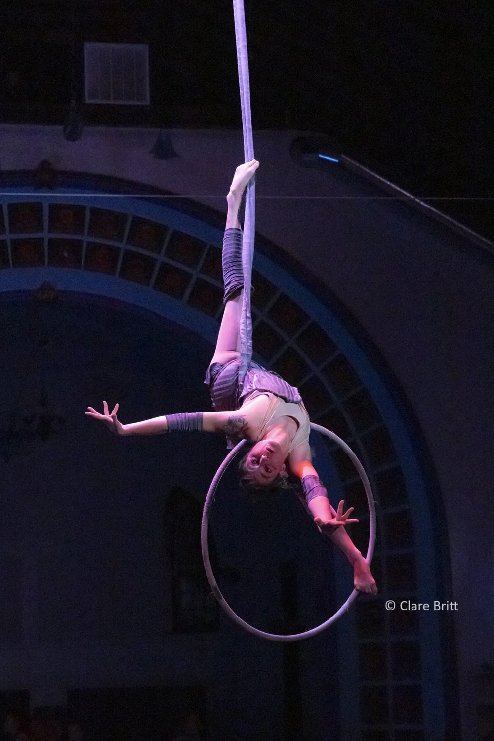 An aerialist performing on an aerial hoop suspended from the ceiling during a circus act, with a dark background and stage lighting. They arch back over the top of the hoop with one leg hooked on the hoop's strap and wear a short grey dress.