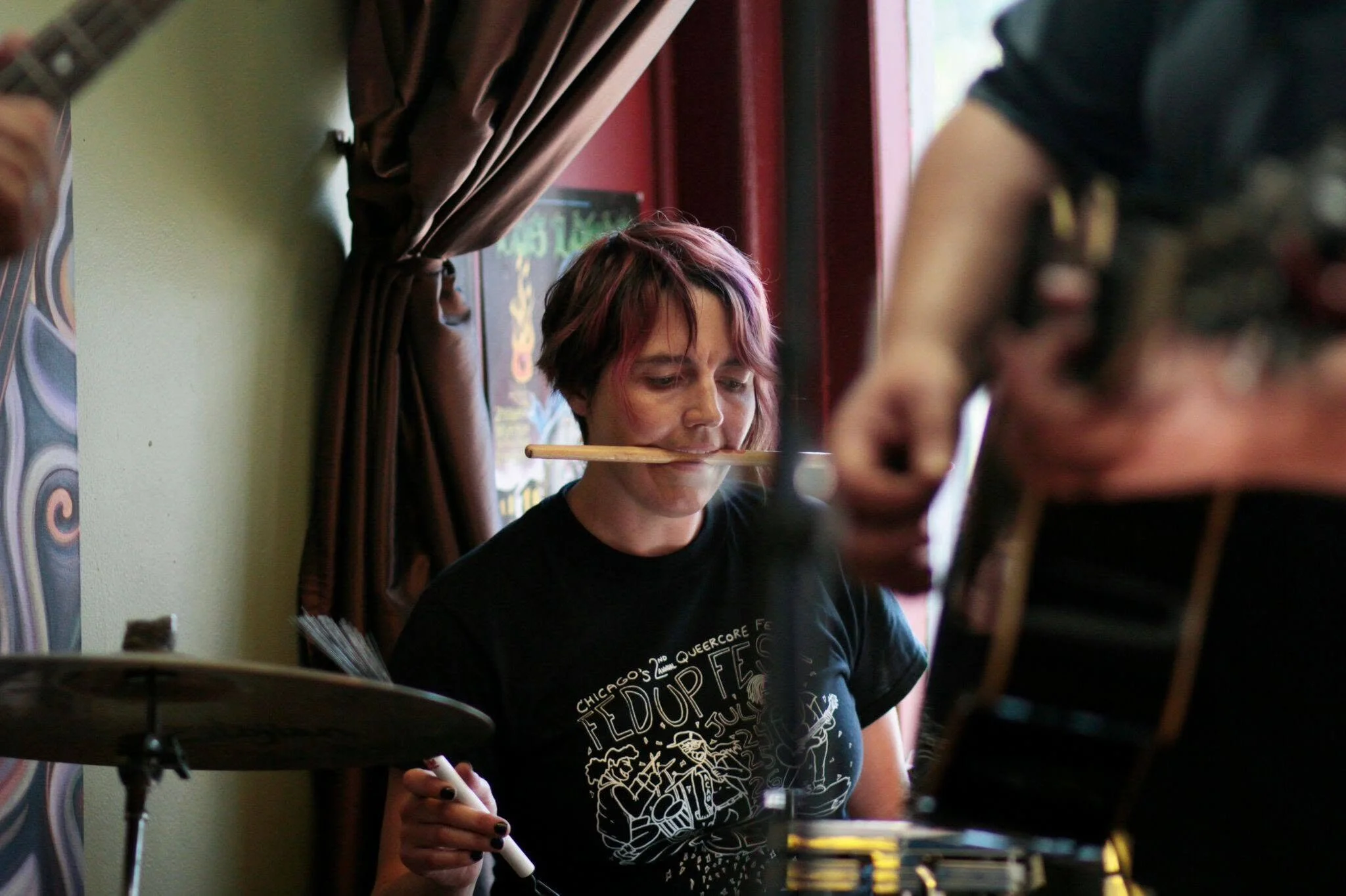 A drummer with brown and purple hair plays drums in the corner of a yellow room.  They have a drumstick in their mouth and are playing with brushes. A guitarist stands to their left and there is a window behind them.