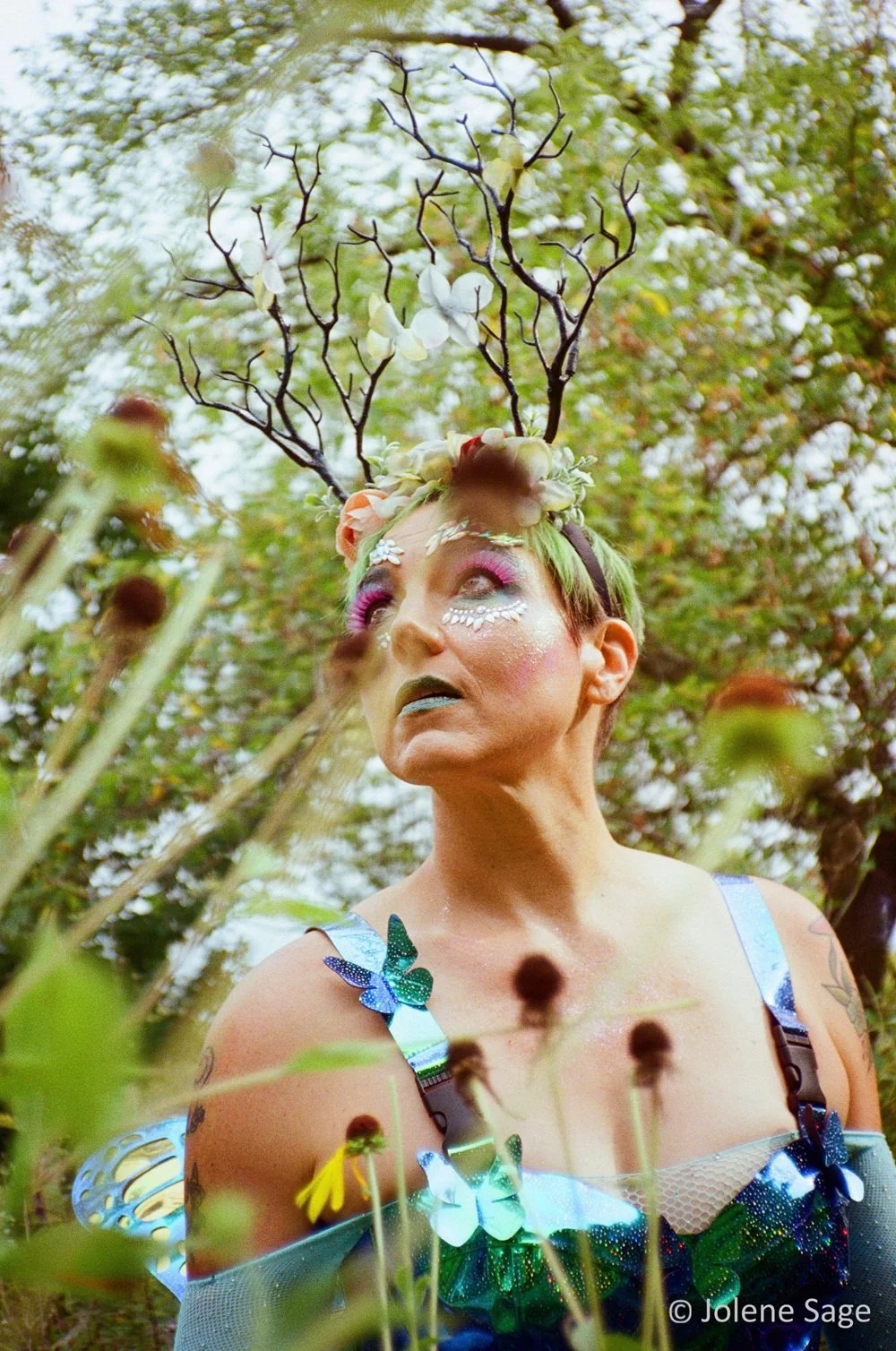 A person with dramatic makeup and colorful costume outdoors, wearing a headpiece with branches and flowers, surrounded by tall plants and trees.