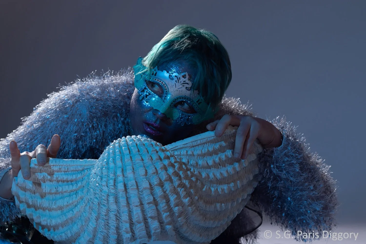 Person wearing a sparkling blue mask with intricate designs, hugging a large artifical seashell, dressed in a fluffy, grey sweater, with short, tousled turquoise hair, against a dark, blurred background.