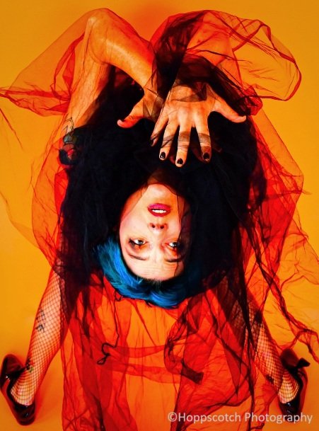 A woman with teal hair wearing a black tulle dress, hangs upside down in a backbend looking at the camera.  They hold their hands near their face.