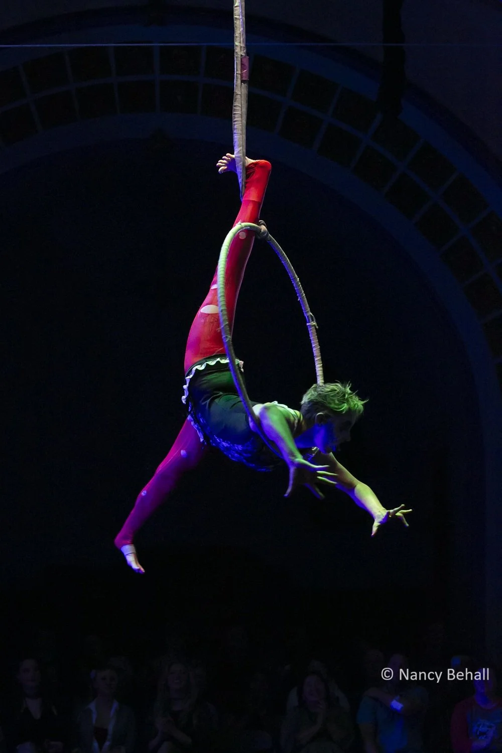 Aerial performer hangs from one leg in an aerial hoop, balanced on their armpits in a split.  They wear a torn black, torn bright red tights and zombie makeup under bright stage lights in a darkened theater.