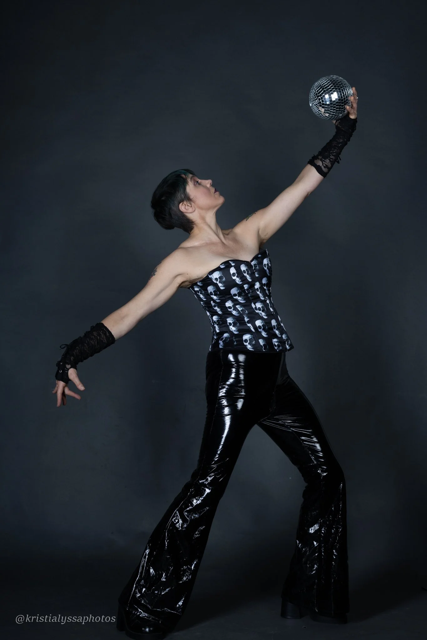 A person with short dark hair wearing a black strapless top with a skull pattern and matching shiny black pants, holding a disco ball above their head with one arm extended upward, posing against a dark background.