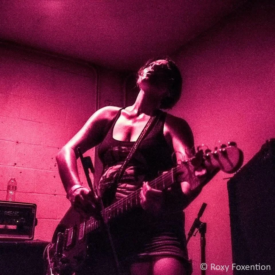 A guitarist wearing a low-cut tank top and denim shorts plays a black electric guitar in a dimly lit room with pink lighting. They glisten with sweat.