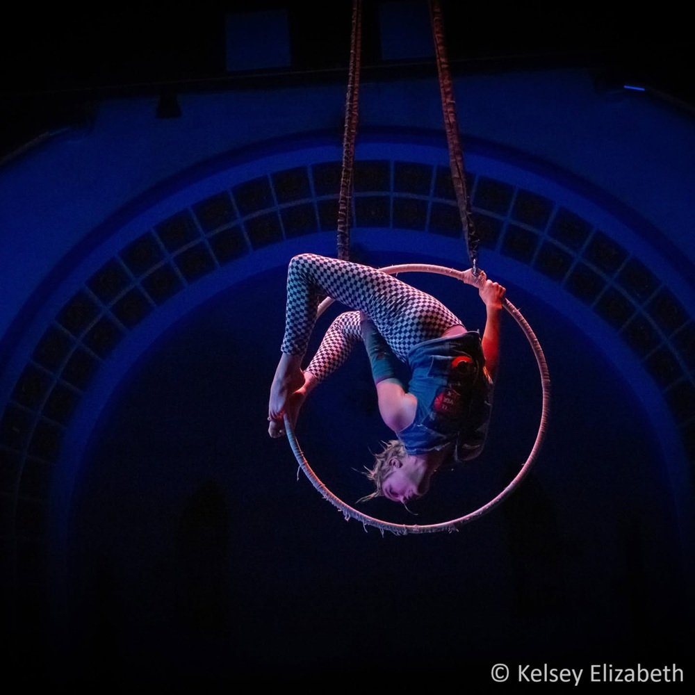 An aerialist with spiky bleached blond hair performs on an aerial hoop in a dark, blue-lit circus theater. They are wearing black and white checkered leggings and a denim vest and hang in a backbend from the top of the hoop.
