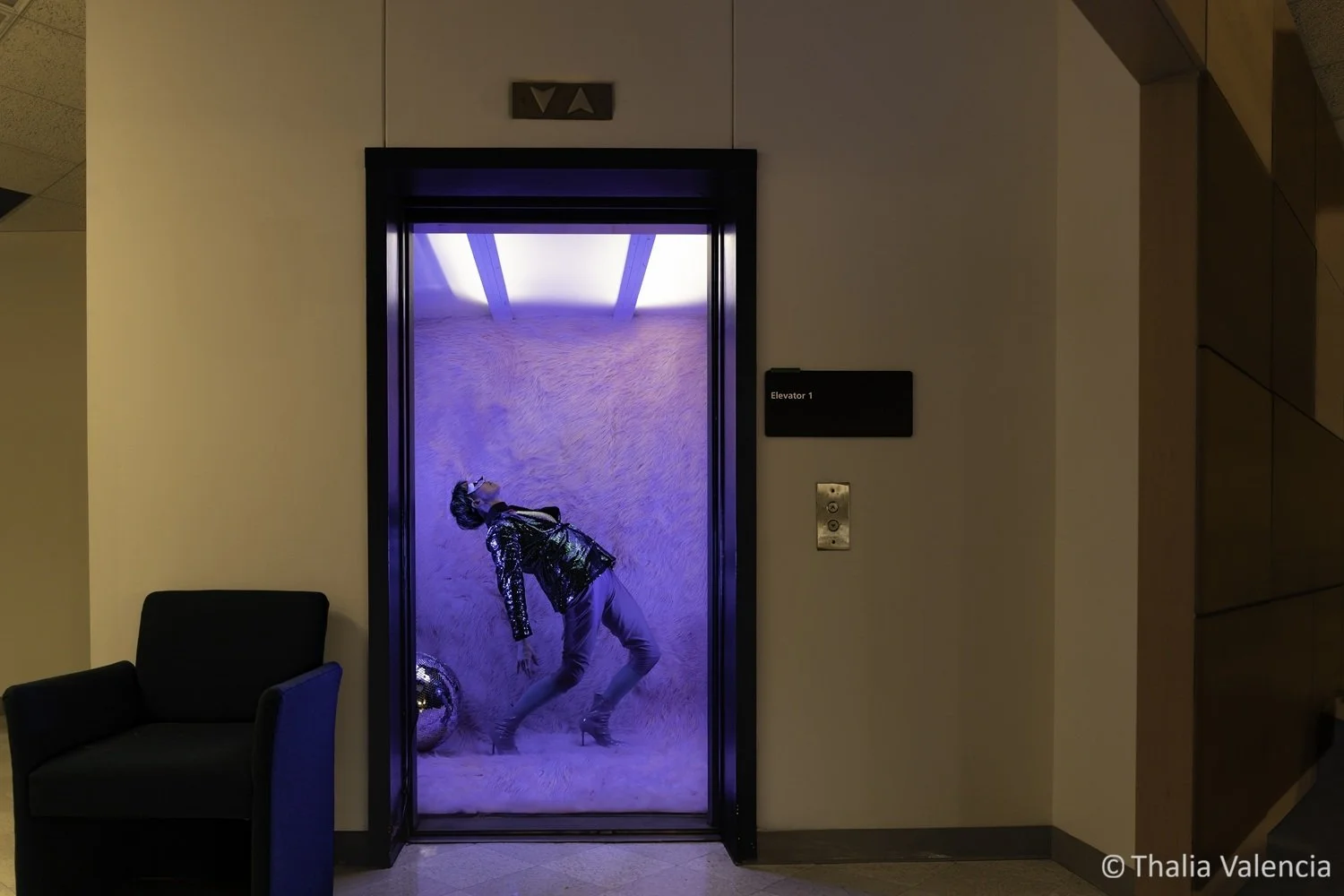 An elevator with its door open, revealing a model in a sparkling jacket and heels, leaning backward against a large disco ball. The elevator is covered with pink fake fur and lit by blue and purple lights.
