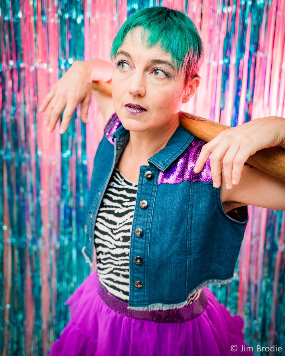 Person with short, teal hair, wearing a denim vest with purple sequin accents, zebra print top, and purple tulle skirt, holding a wooden baseball bat on shoulders, standing in front of glittery, colorful backdrop.