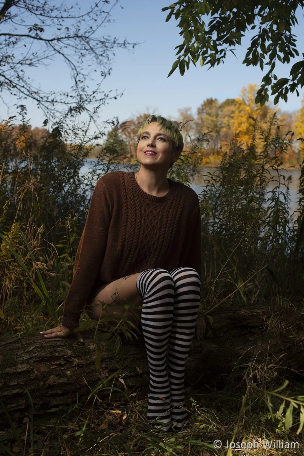 A person with short yellow-green hair sitting on a fallen log in a natural outdoor setting with trees and a lake in the background, wearing a brown sweater and black-and-white striped tights, gazing upwards with a smile.