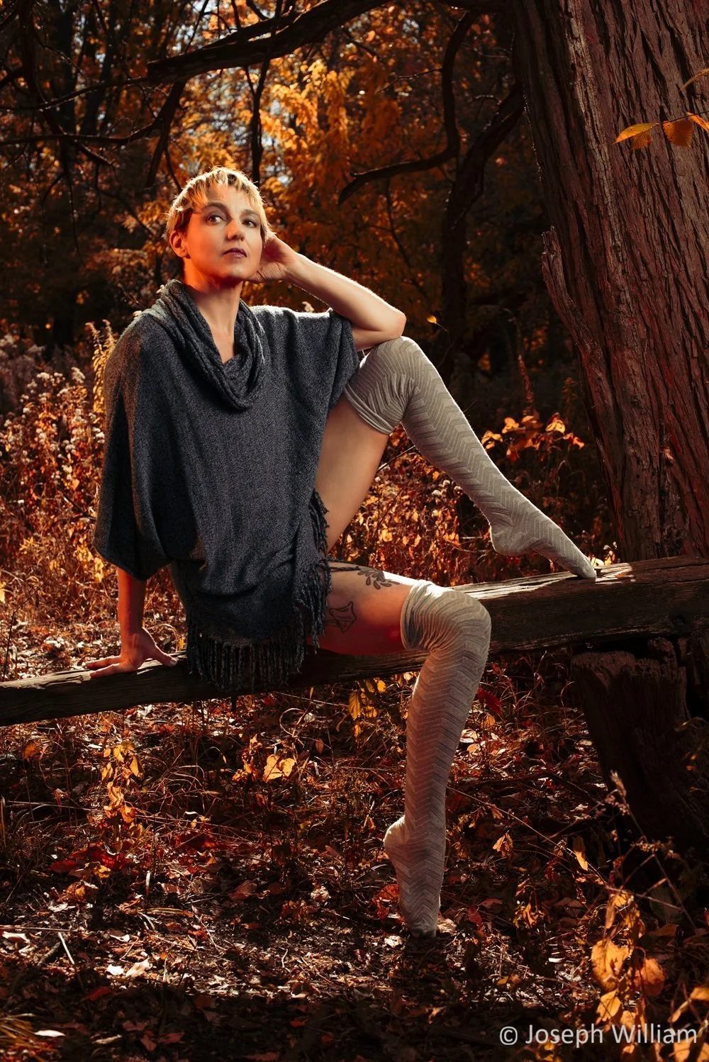A person with short blond hair sitting on a wooden rail in an autumn forest, wearing a blue sweater, striped thigh-high grey patterned socks.  They smile and look upward, surrounded by sunlight.