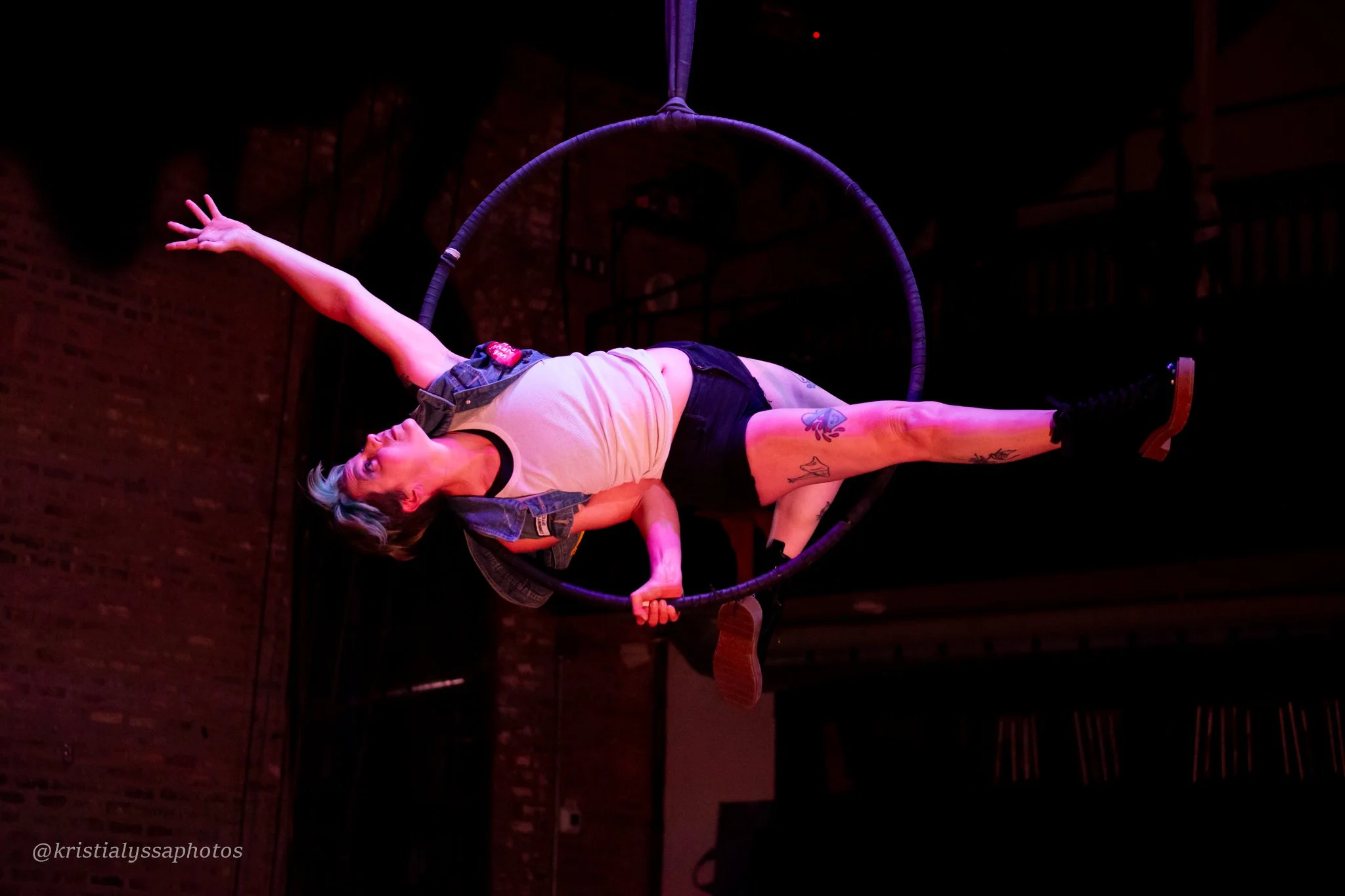 An aerial performer in a white tank top, black shorts and denim vest balances in black aerial hoop in a dimly lit venue with brick walls. They are laying propped on one elbow with one arm extended upward and both legs extended out.