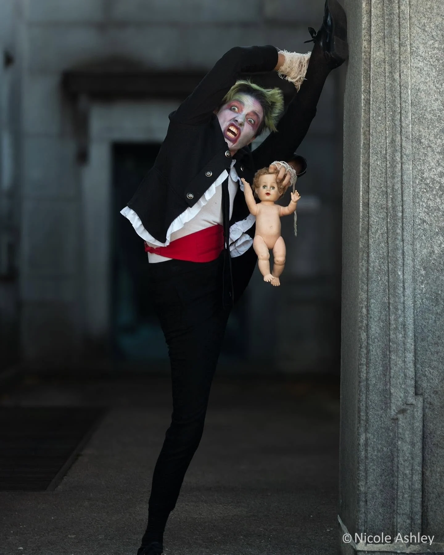 Person with zombie makeup and green hair, dressed in black and white formal wear and a red cumberbund, holding a doll, standing in the splits against a concrete wall in a dark outdoor setting. They have a dramatic expression and look menacing.