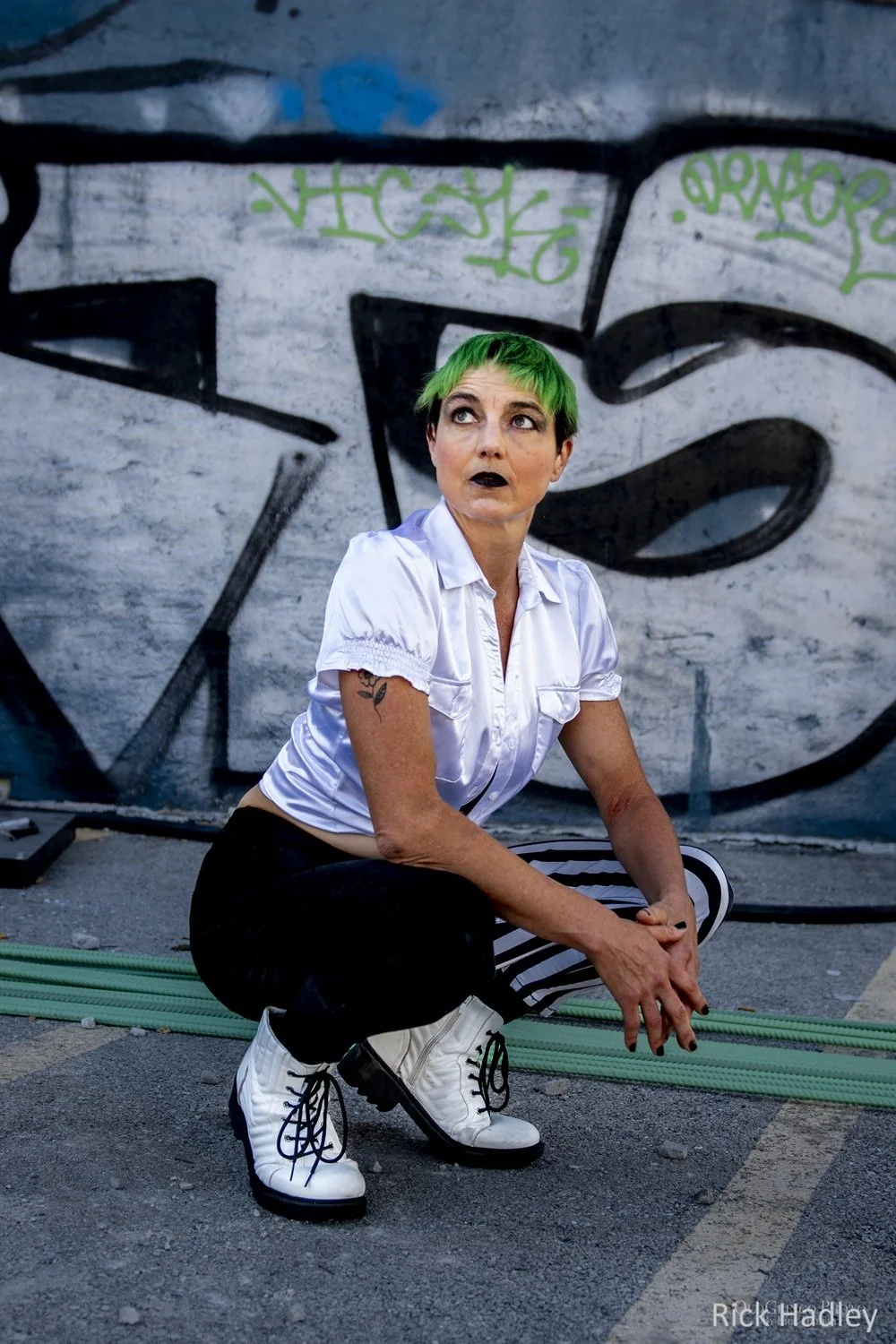 A model with short green hair, wearing a white blouse, striped black and white pants, and white boots, crouches in front of a graffiti-covered wall.