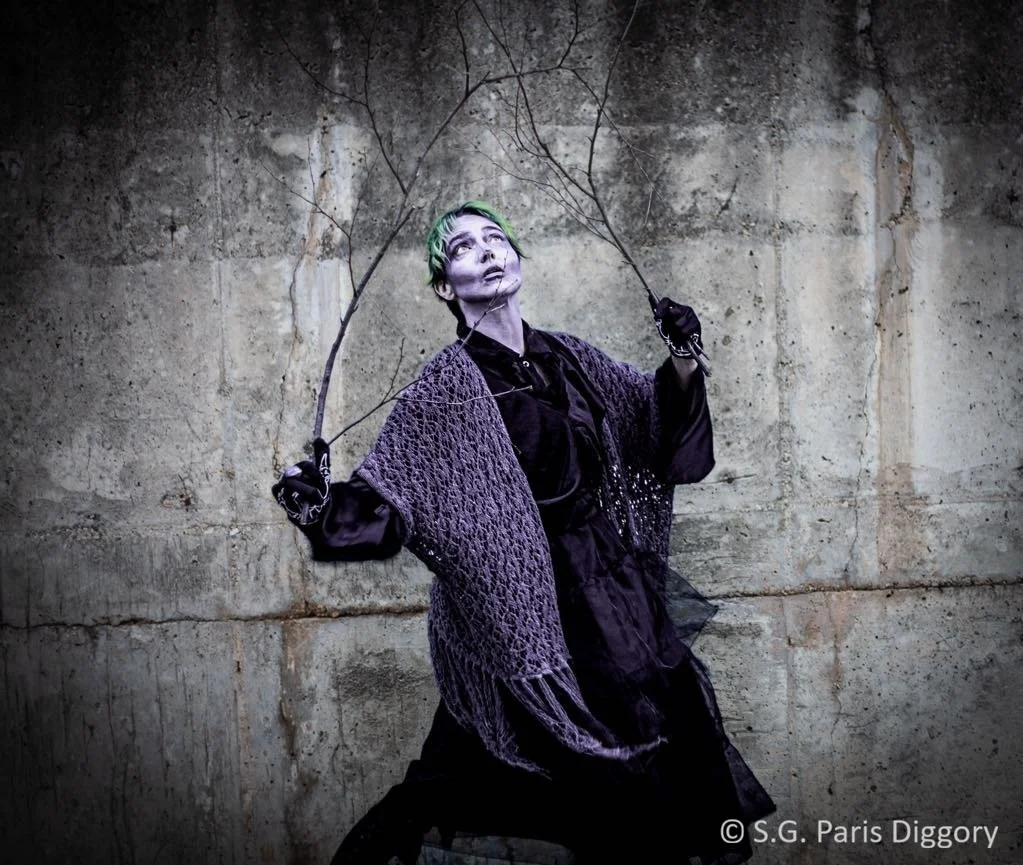 Person with green hair, gothic makeup, and dark clothing stands against a concrete wall, holding two thin branches.