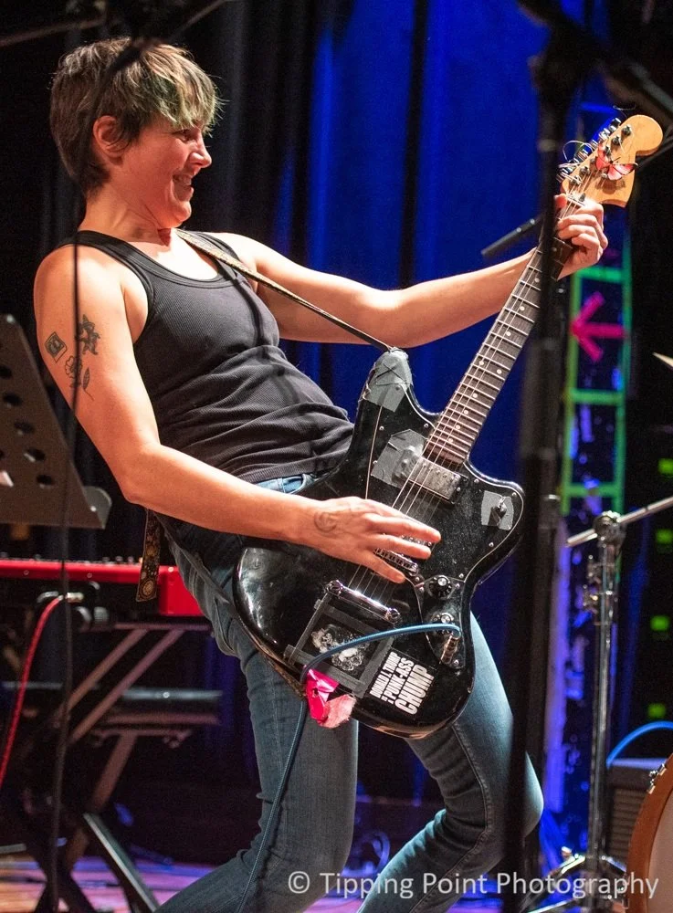 A non-binary guitarist with short brown hair plays a low slung black electric guitar on stage, smiling and leaning back, with stage equipment and colorful lights in the background.