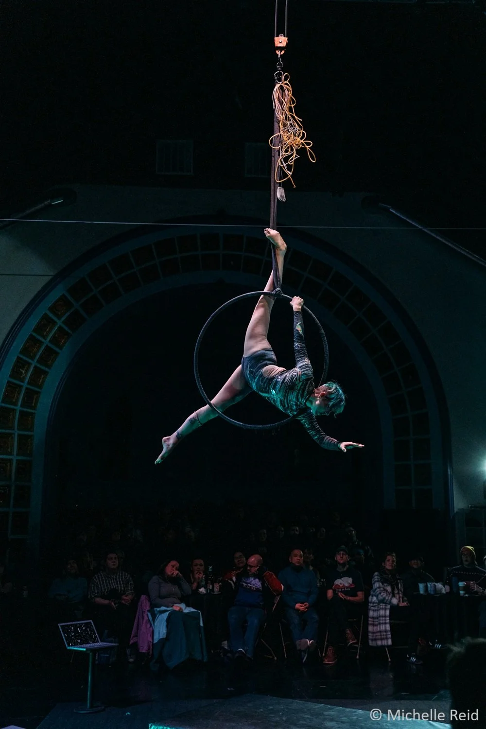 An aerialist performs on an aerial hoop hanging from the ceiling in front of an audience in a dark theater. One of their legs is hooked on the rope above the hoop and the other stretches towards the floor in a split.  Their hands reach out wide.