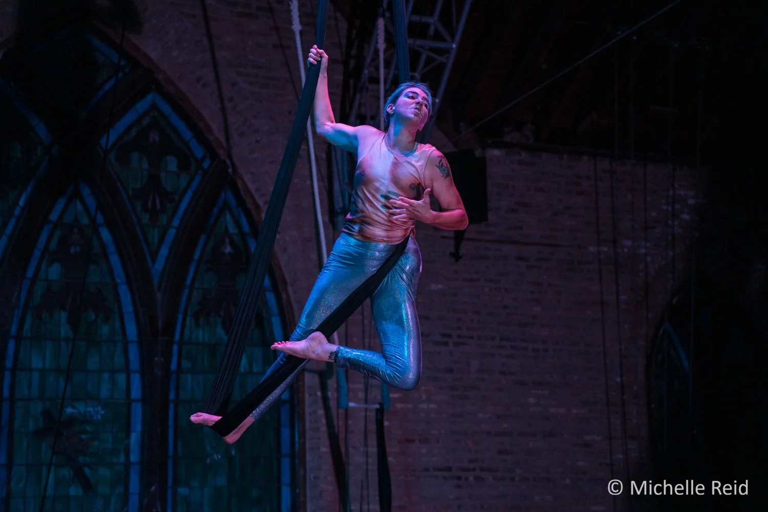 A aerialist in drag king makeup performs on a black aerial sling in a circus theater setting.  They wear a shirt patterned like a male chest and shiny silver leggings, holding the sling with one hand and touching their chest with the other.  