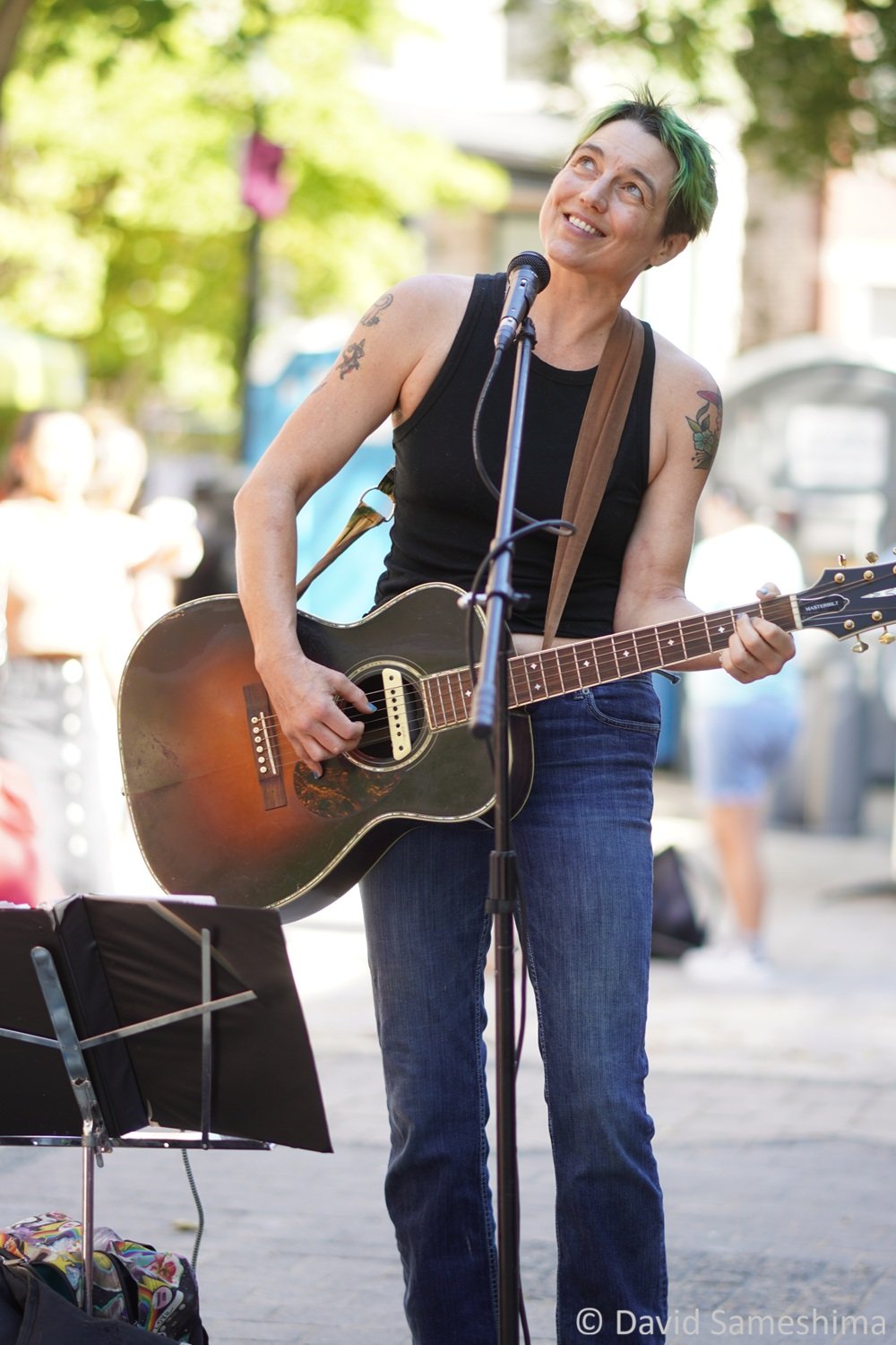 A non-binary musician with short green hair wears a black sleeveless top and blue jeans playing an acoustic guitar on a city plaza during the day. Green trees and spectators are visible behind them.