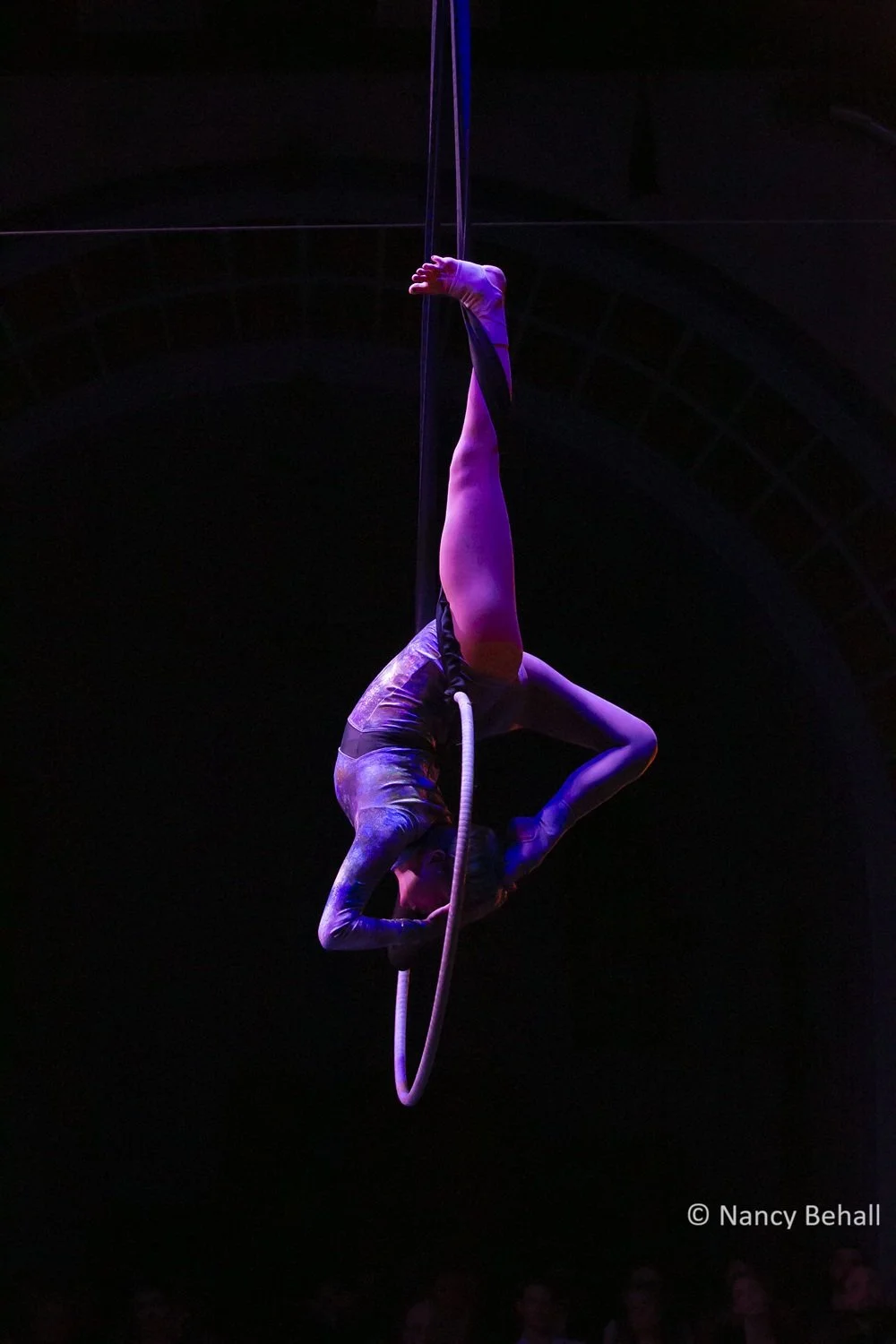 Aerial performer hanging upside down from a vertical ring, illuminated with purple and pink lighting against a dark background. They have their foot pulled to their head from behind and are wearing a sparkly purple and black leotard.
