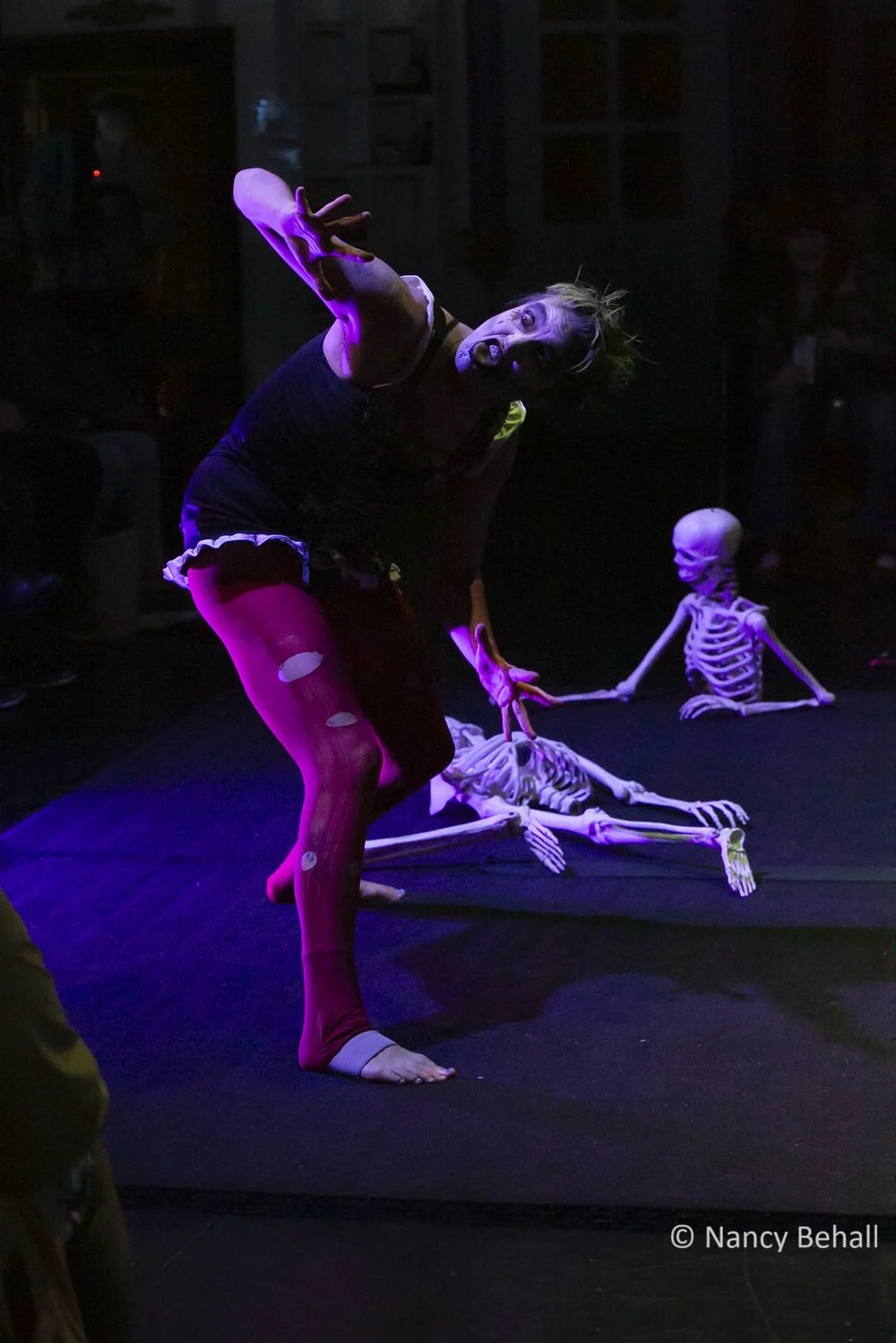 Performer in black dress, ripped red tights and zombie makeup poses with skeleton props on stage in a dark theater.