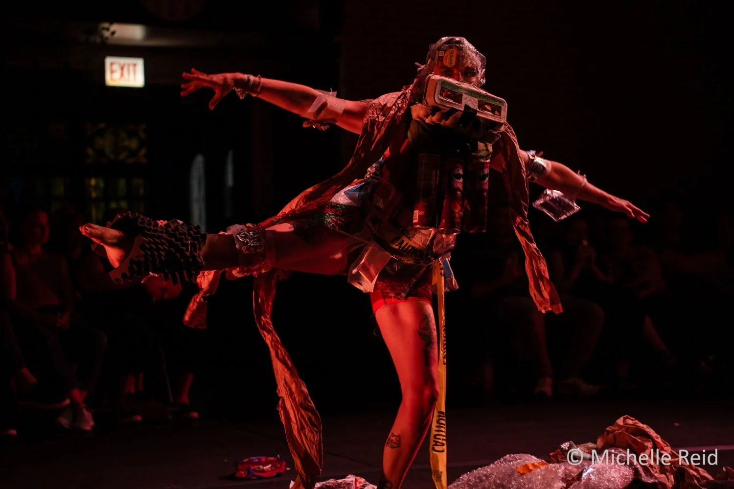 Performer balancing on one leg on stage, wearing an elaborate costume made of junk like cardboard, plastic, and tape, with arms outstretched. Audience members observe in the background, and dramatic red lighting highlights the scene.