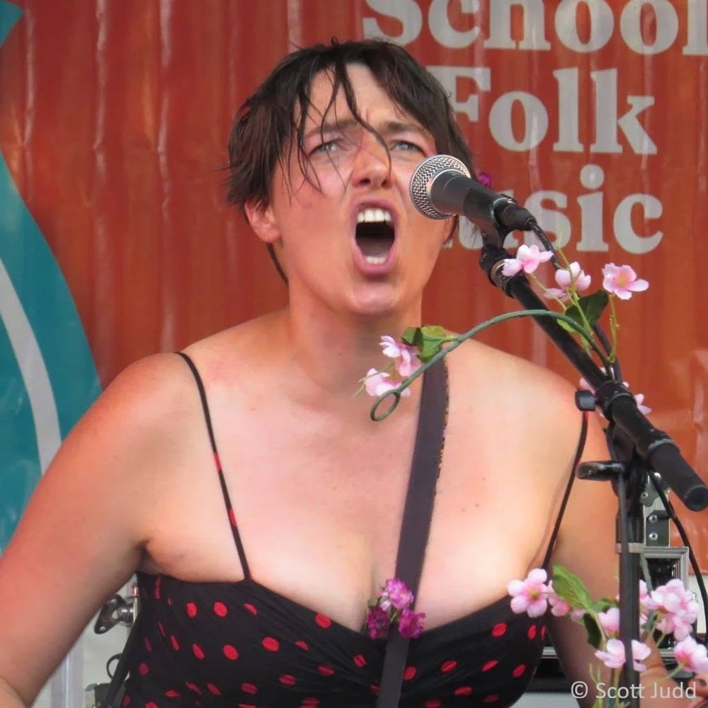 A vocalist sings passionately into a microphone on an outdoor stage, wearing a black dress with red polka dots, with short brown hair and pink flowers attached to the microphone stand. An orange banner for a folk music festival is behind them.