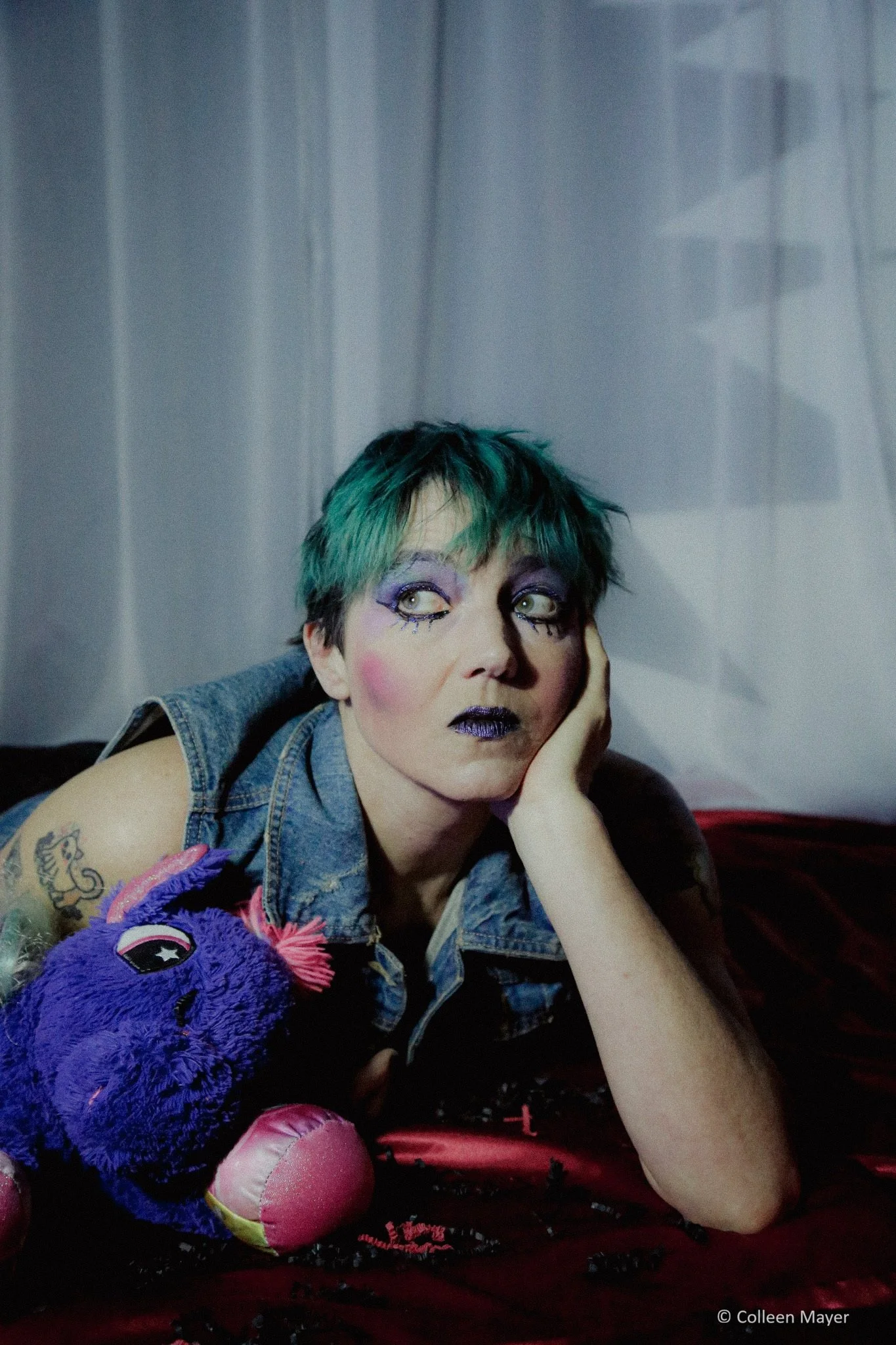 A non-binary model with green hair and theatrical pink, purple and blue makeup stares out beyond the camera.  They are wearing a denim vest and holding a purple stuffed animal.