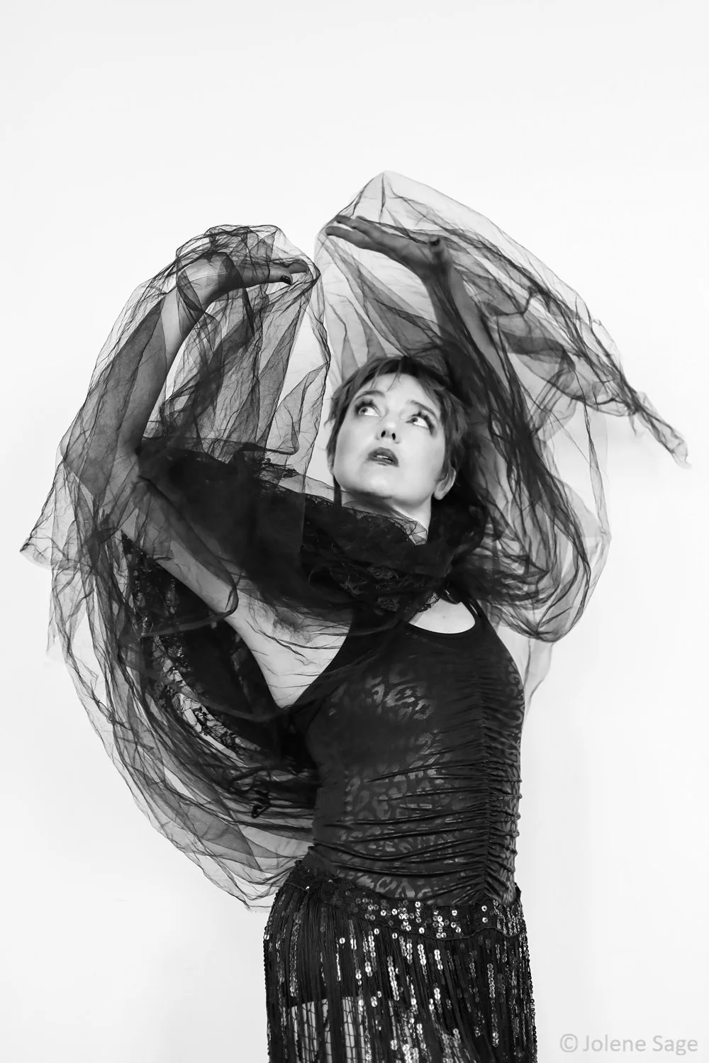 A person with short dark hair wearing a black, sleeveless, lace top and sequined skirt, holding sheer tulle fabric above her head in an arc against a plain background, looking upwards.