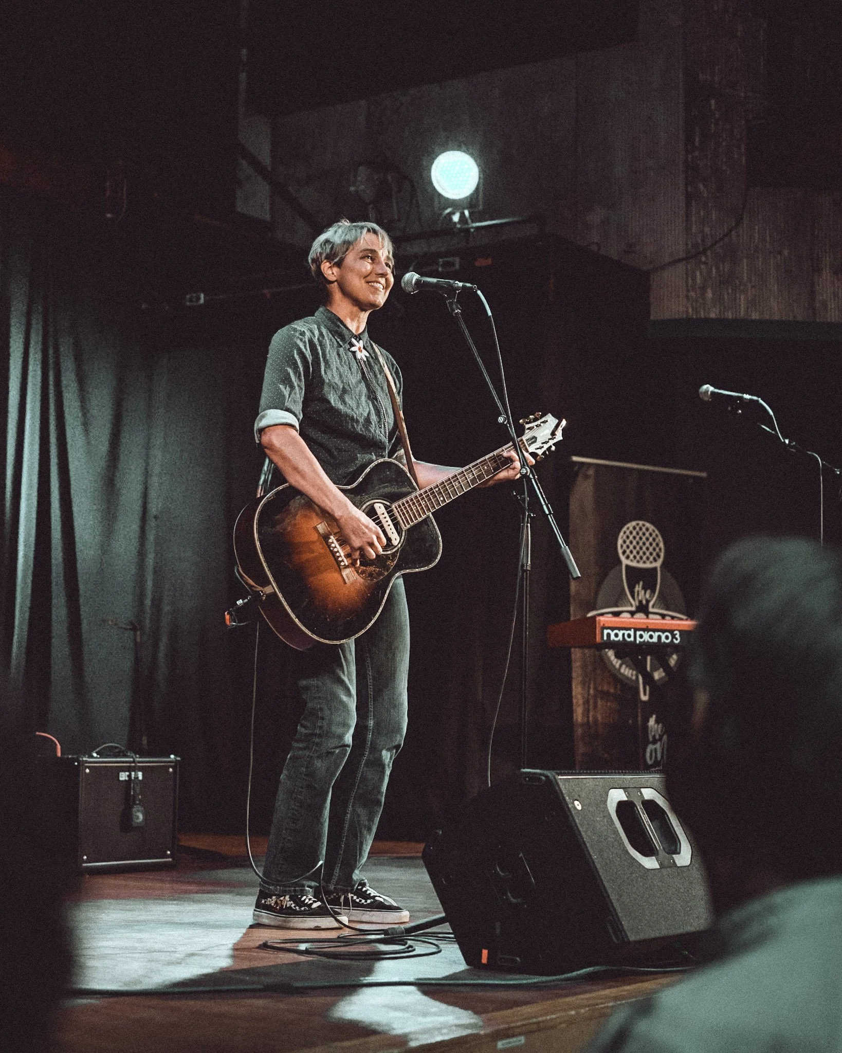 A young person with short, grayish hair is playing an acoustic guitar on stage, smiling, and singing into a microphone. They are wearing a dark, patterned shirt and jeans with sneakers. The stage has musical equipment, including a keyboard and speakers, with dark curtains and a spotlight in the background.
