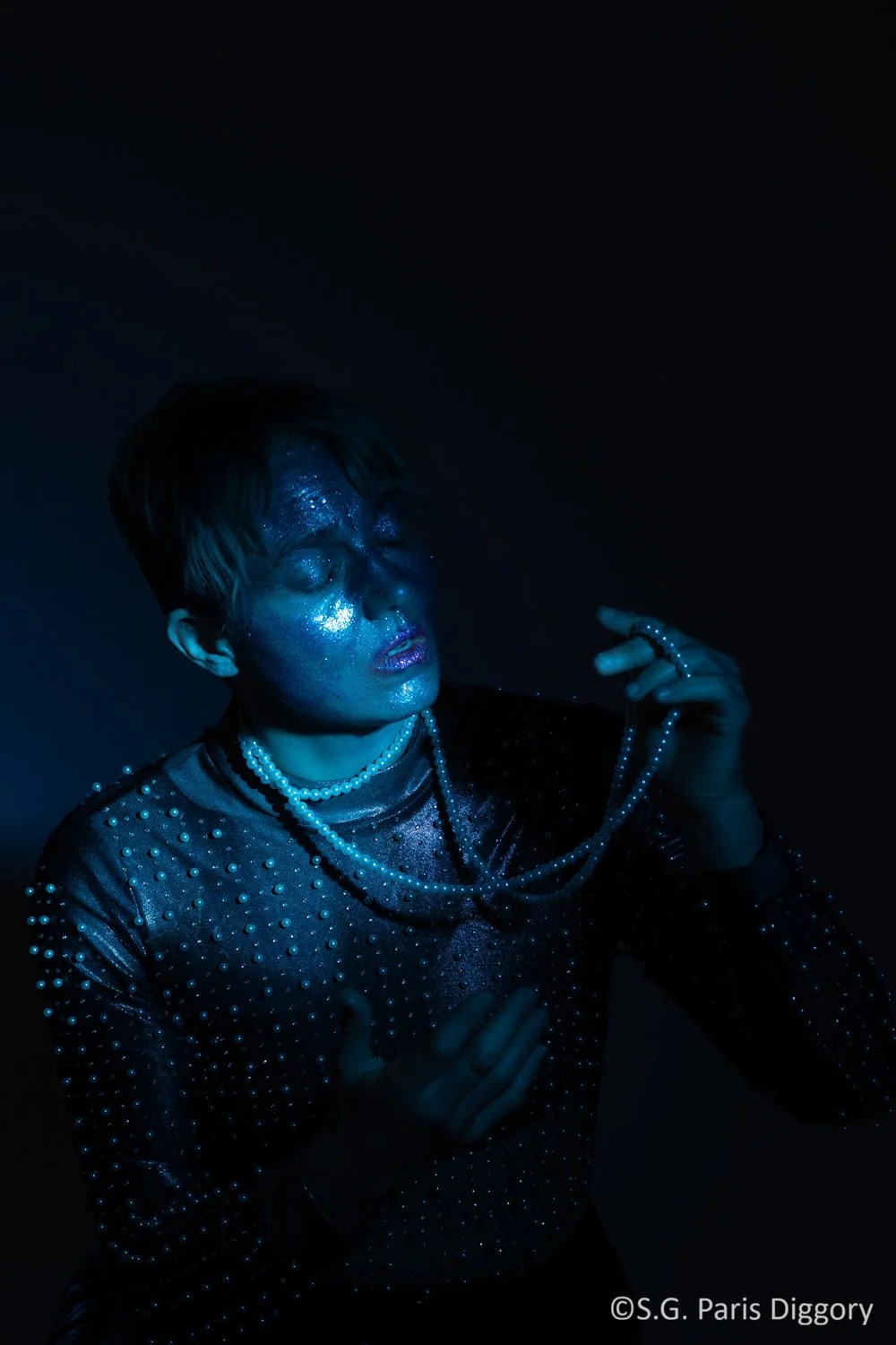 Person with short hair, covered in blue glitter and pearls, posing with eyes closed in darkness, illuminated by blue lighting.
