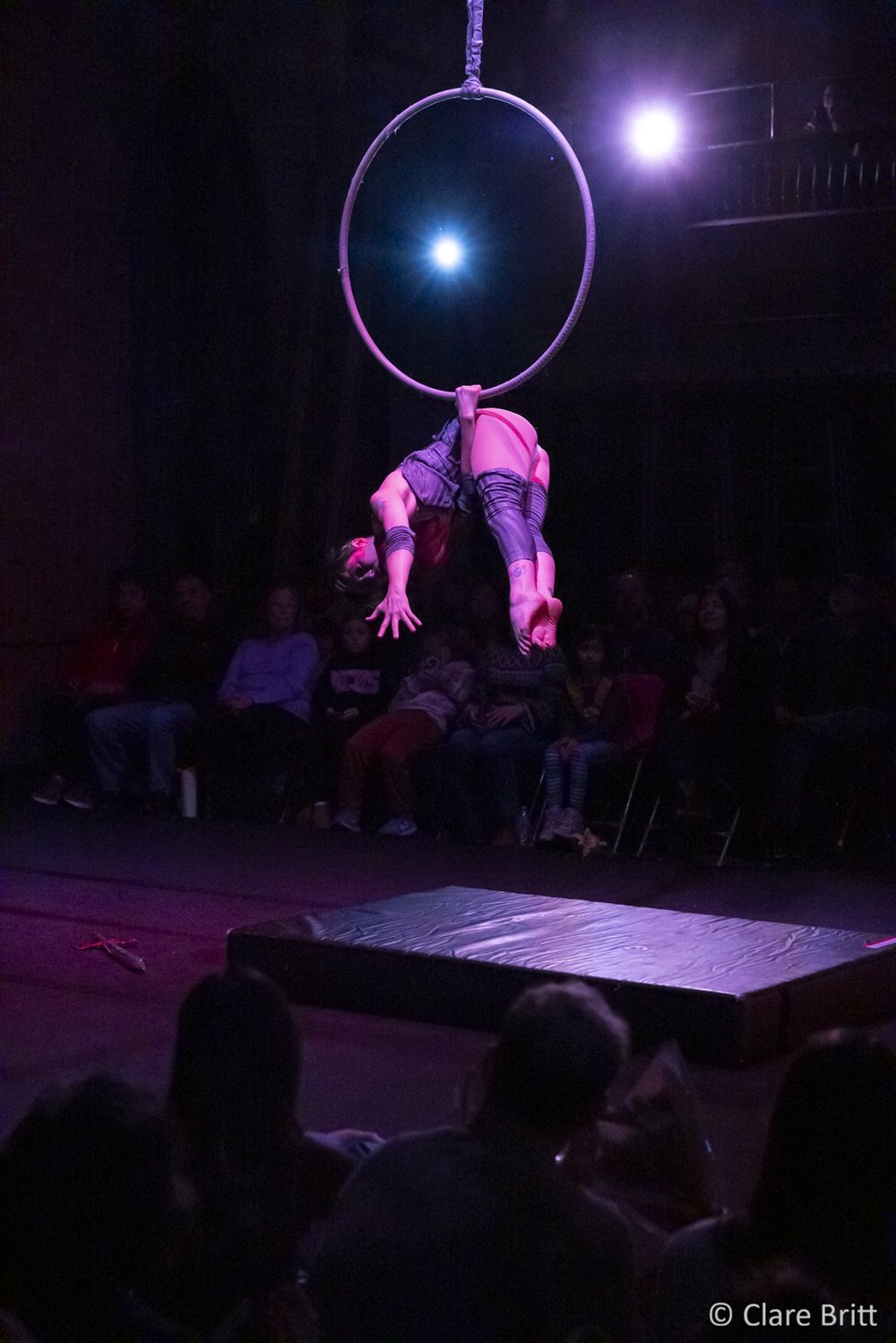 An aerialist hangs upside down from one arm, with their other arm and legs reaching towards the floor.  They are wearing a short grey dress and leg sleeves and an audience watches in the dark behind them.