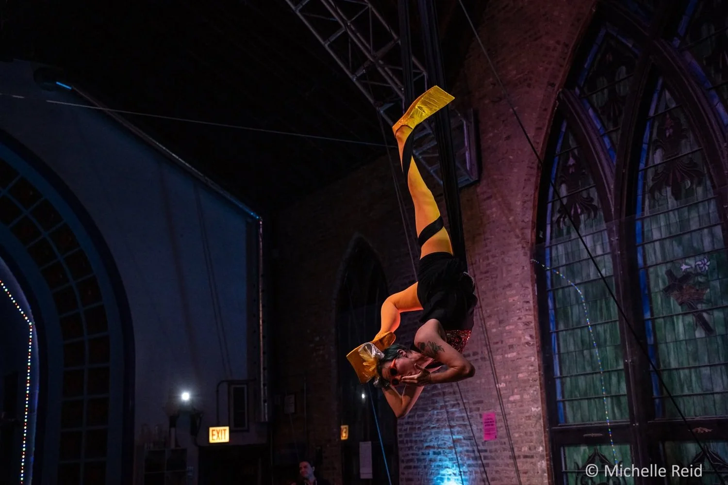 An aerialist performing an aerial sling act hangs upside down in a dimly lit venue with stained glass windows and brick walls.  They are wearing yellow tights, yellow swim fins, a black cheerleading costume and  orange sunglasses.