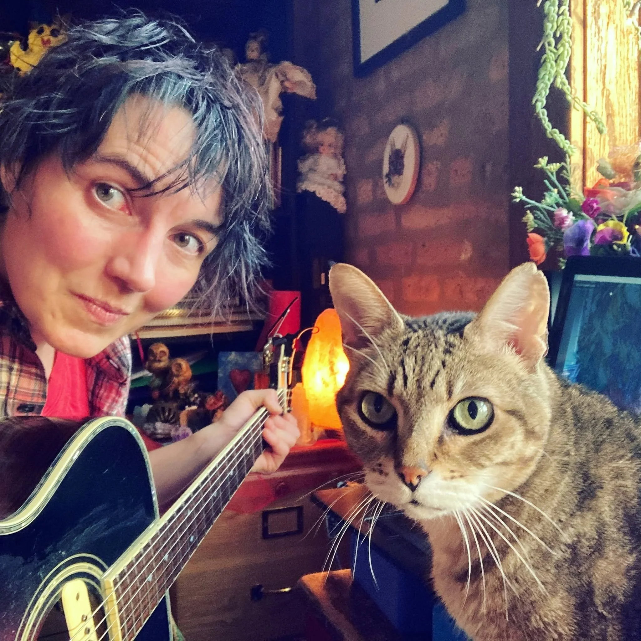 A non-binary person with green hair holds an acoustic guitar and leans towards a tabby cat sitting on a short red desk.  A brick wall and plants are behind them.
