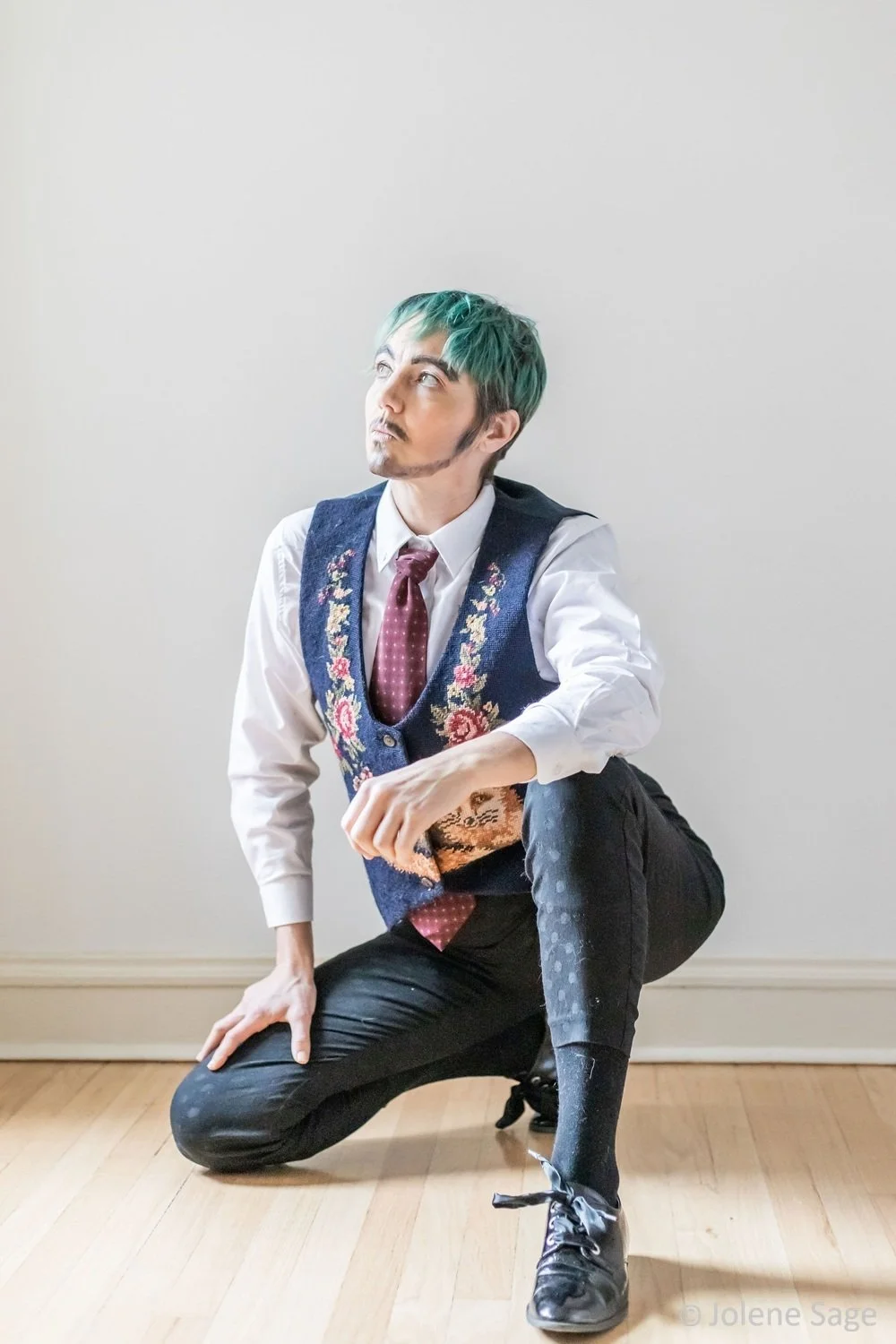 Person with short teal hair squatting on a hardwood floor against a plain light-colored wall, wearing a white shirt, a floral vest, a maroon tie, dark pants, and black shoes.  They have painted on facial hair and are illuminated by sunlight.