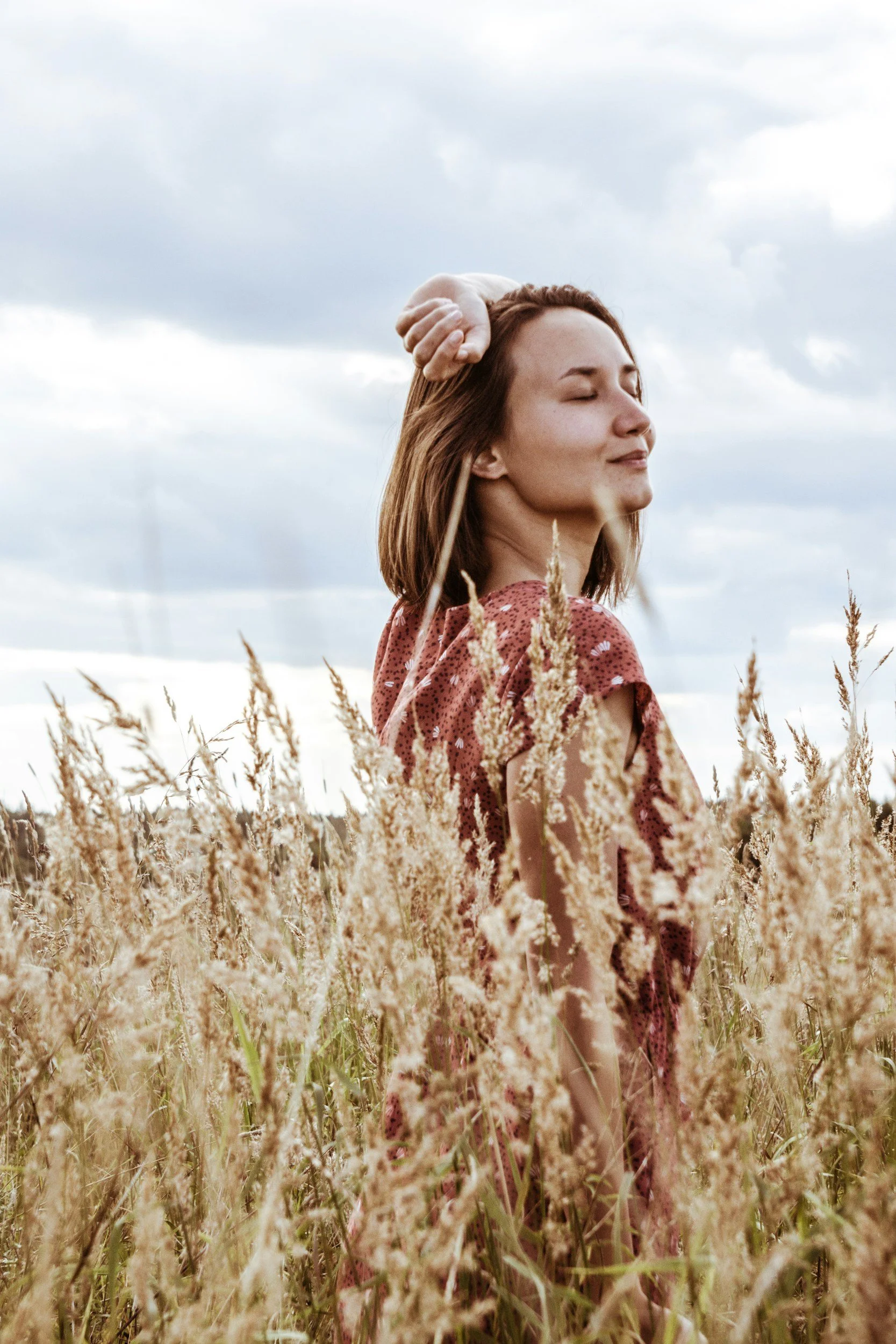 A young woman with closed eyes and a slight smile standing in a field of tall, dried grass under a cloudy sky, with her right arm raised and hand resting on her head.
