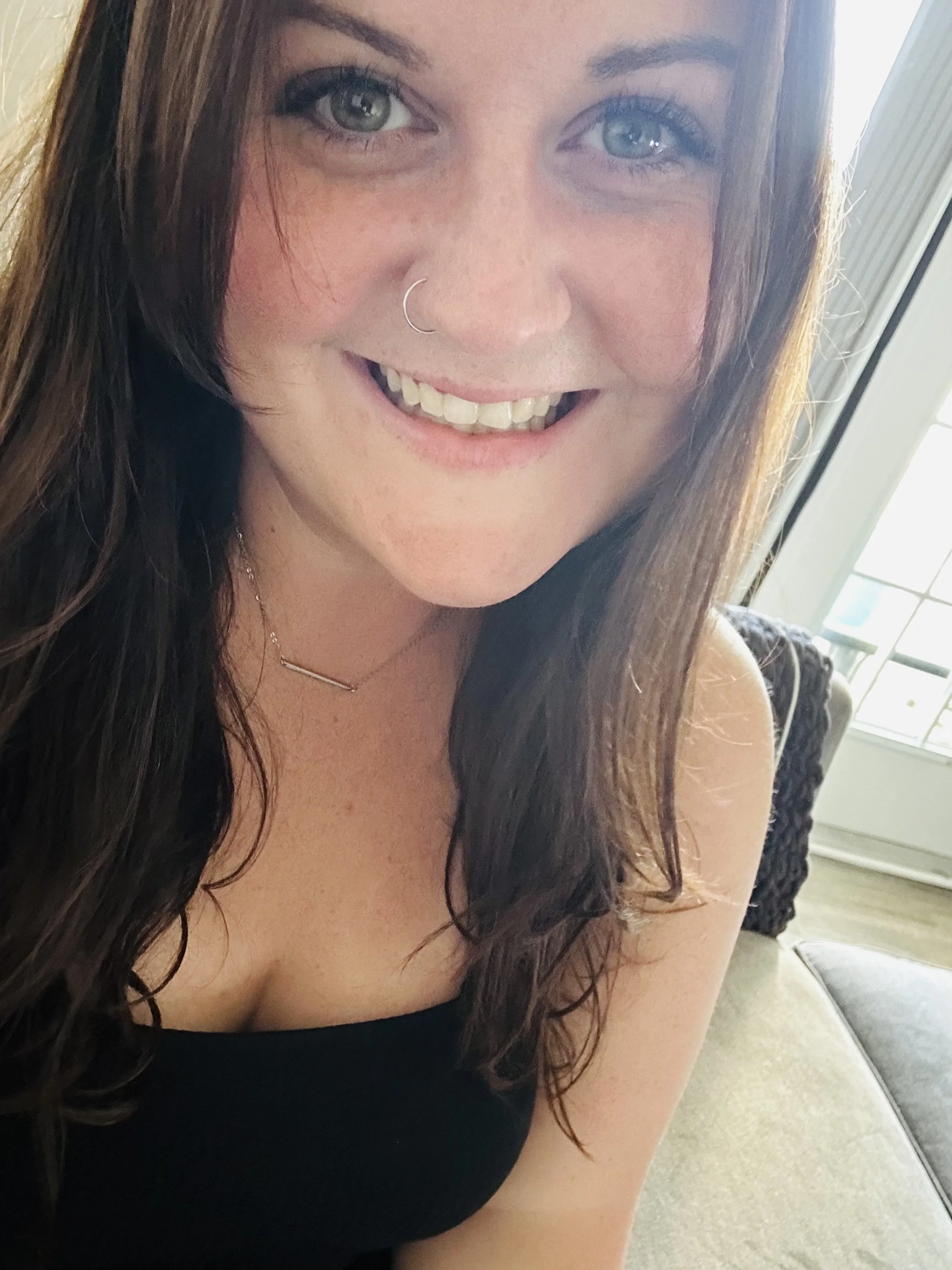 Close-up of a woman with long dark hair, blue eyes, and a nose ring smiling, near a window with sunlight streaming in.