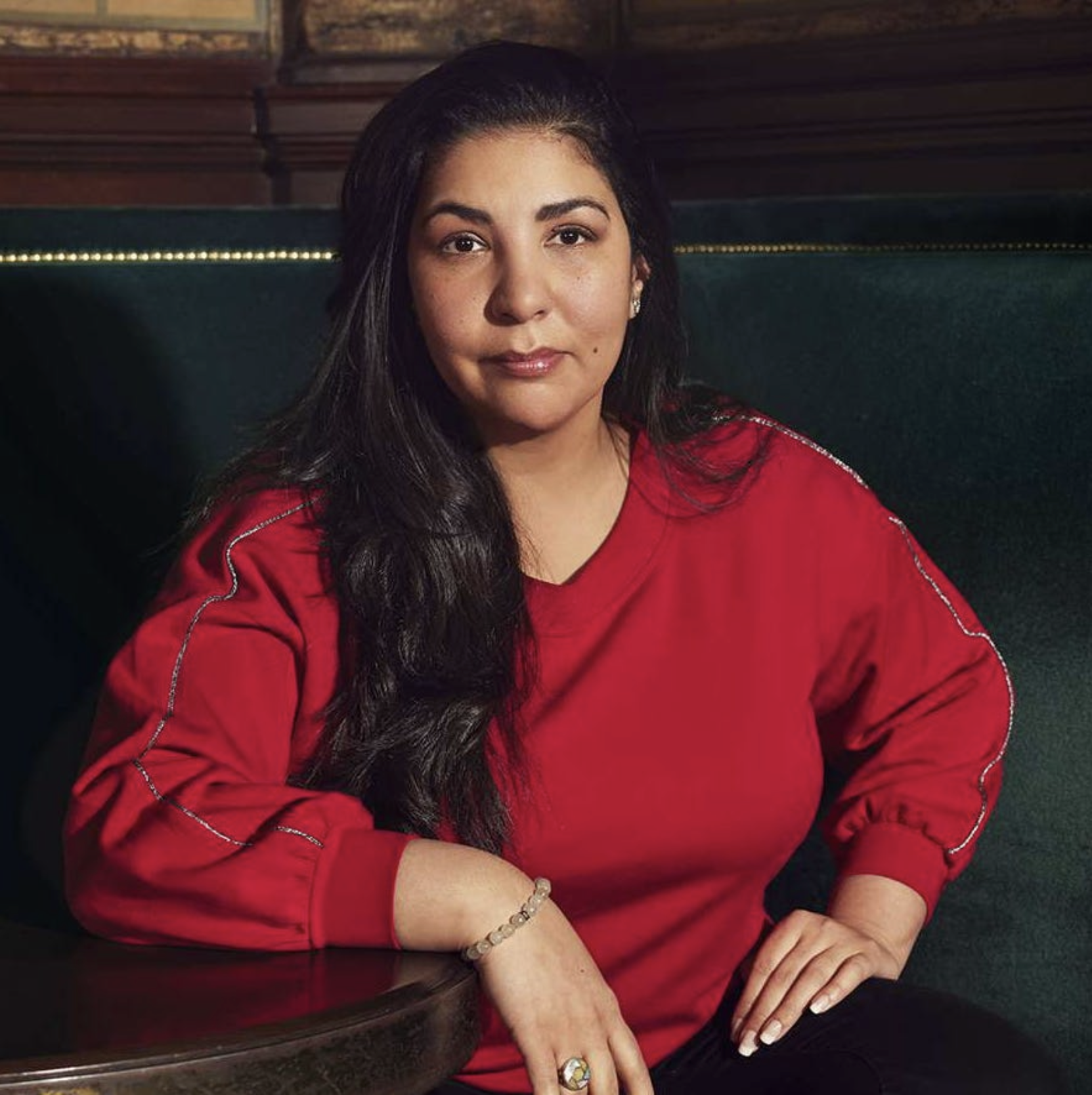 A woman with dark hair, wearing a red shirt, sitting at a dark table in a dimly lit setting with wooden walls behind her.