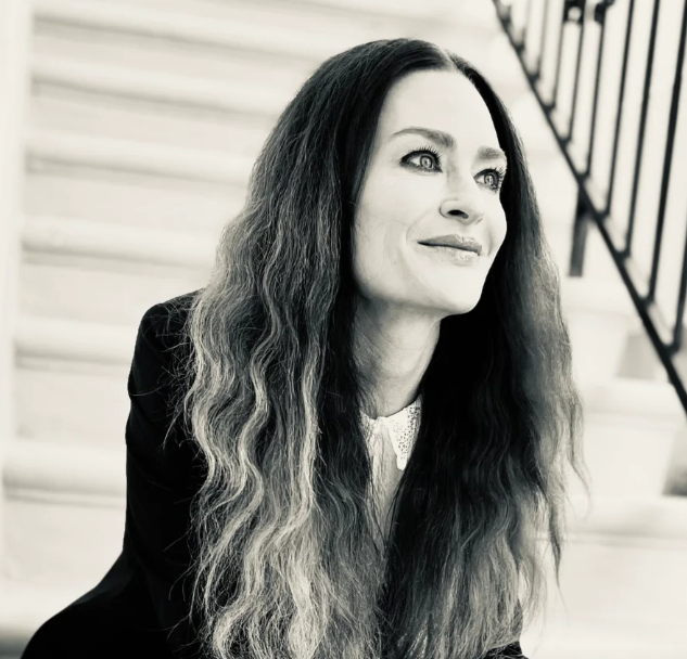 A woman with long, wavy hair, wearing a dark blazer and light-colored blouse, sitting on stairs and looking to the side, smiling slightly.