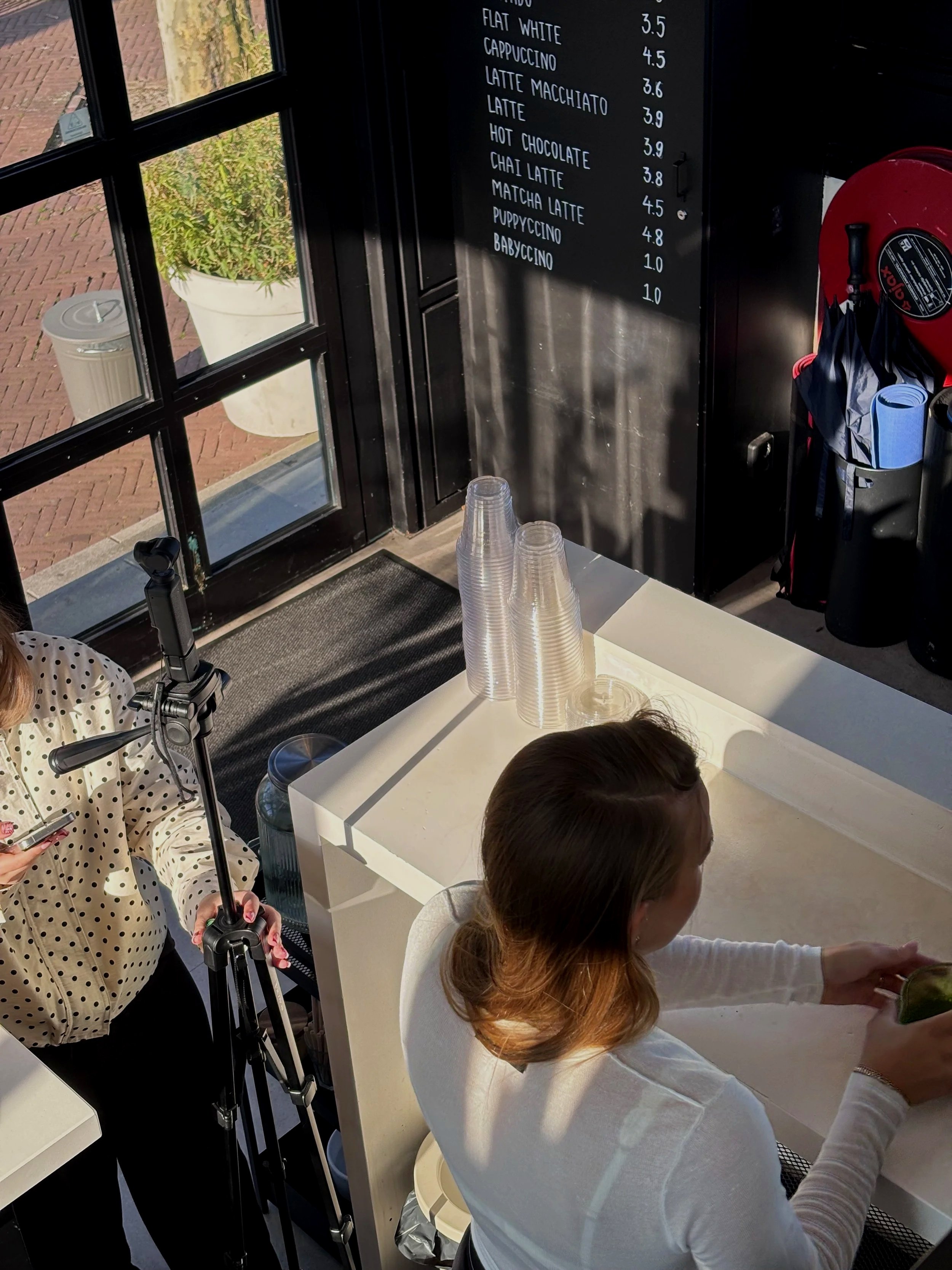 Inside a cafe with a black menu board listing coffee drinks and prices on the wall in the background. A woman with reddish-brown hair is sitting at a white table with a girl in a white sweater, both reaching for something. A person in a polka dot shirt is holding camera equipment near the window, which shows a flower pot outside.