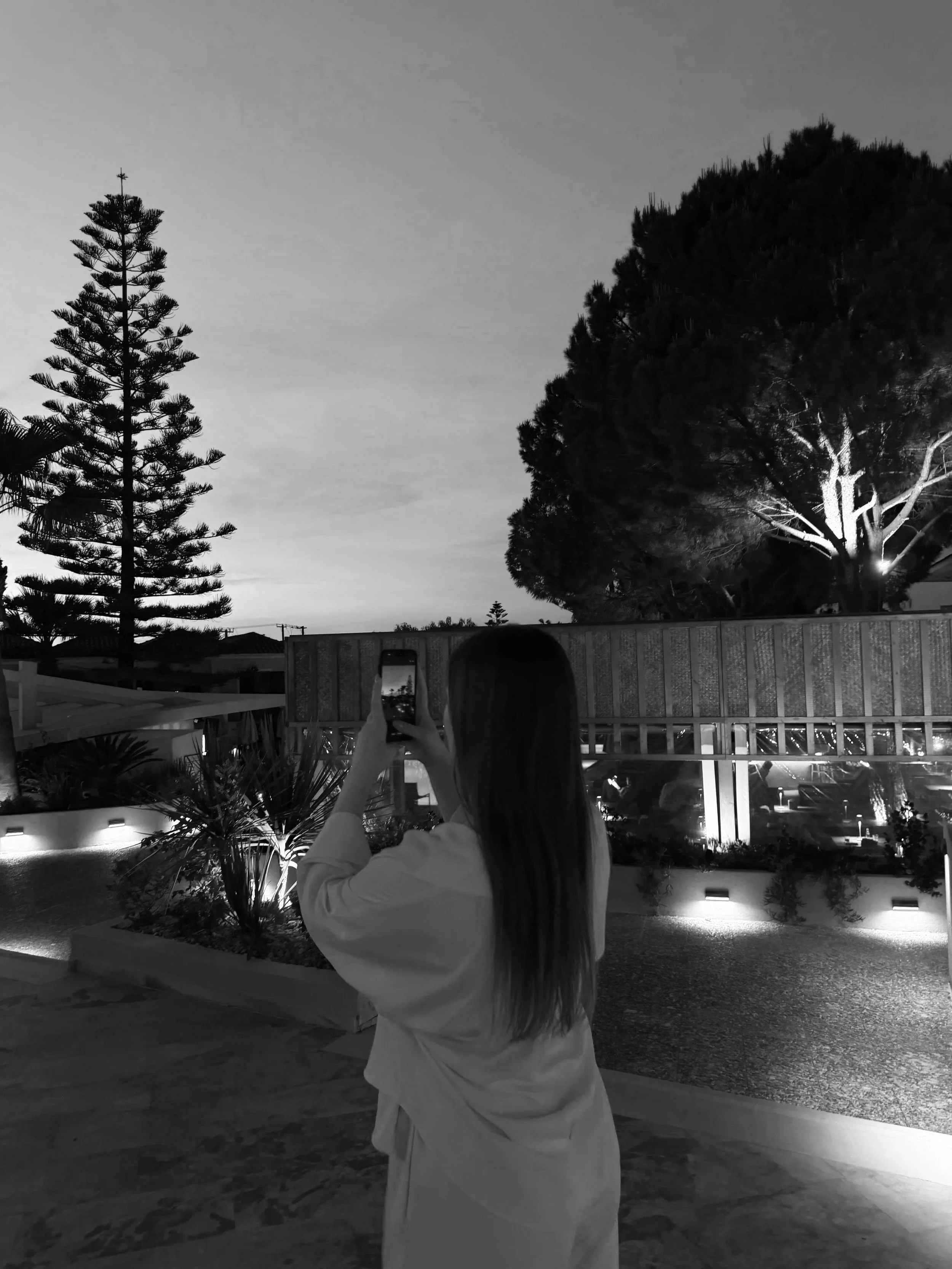 A woman with long hair wearing a light-colored jacket is taking a photo of the night sky with her smartphone. There are large trees and a building with lit windows in the background, under a partly cloudy sky.