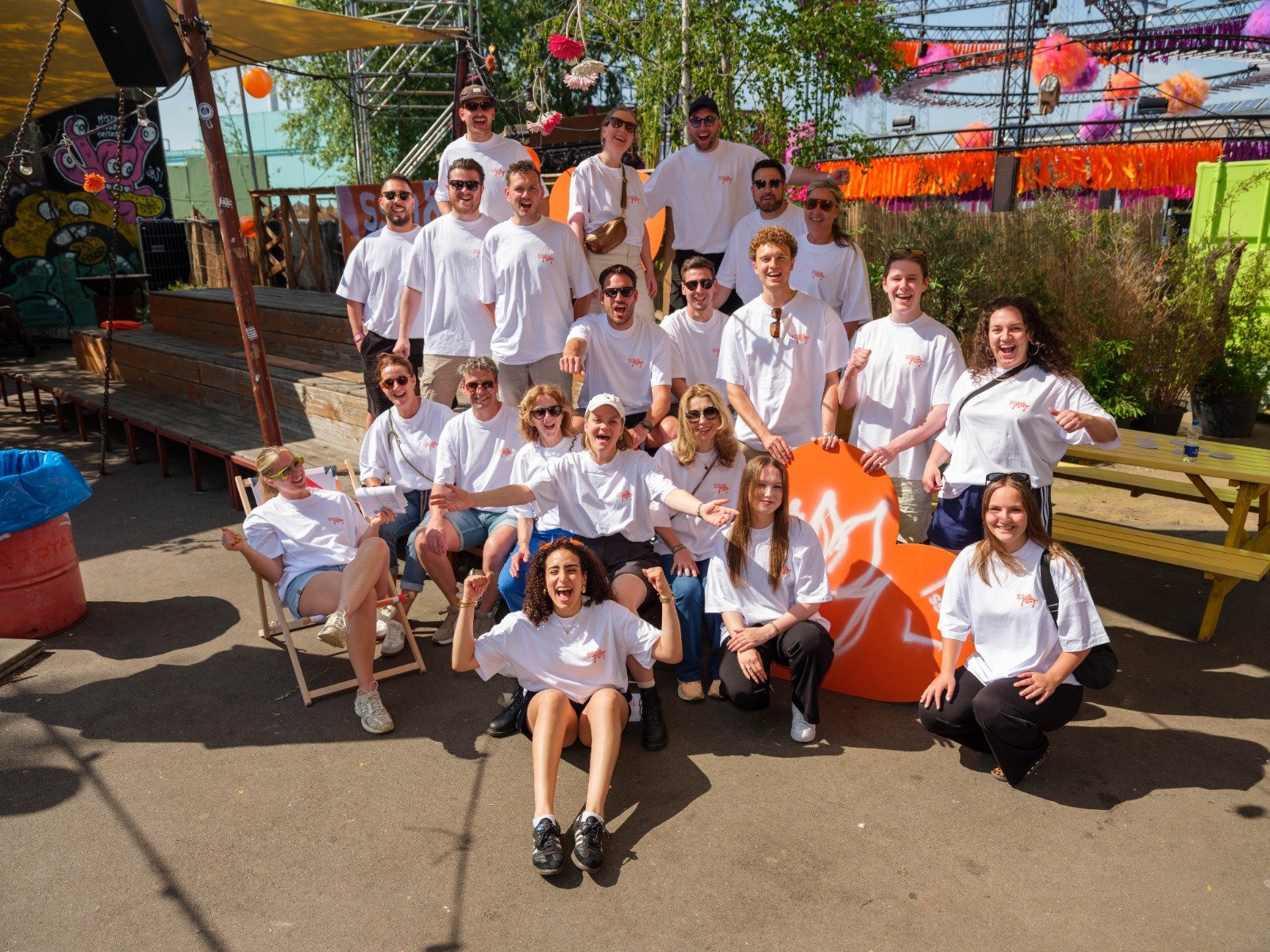 A large group of people posing outdoors at a festival or event, many wearing white T-shirts, some sitting, some standing, with colorful decorations and a roller coaster in the background.