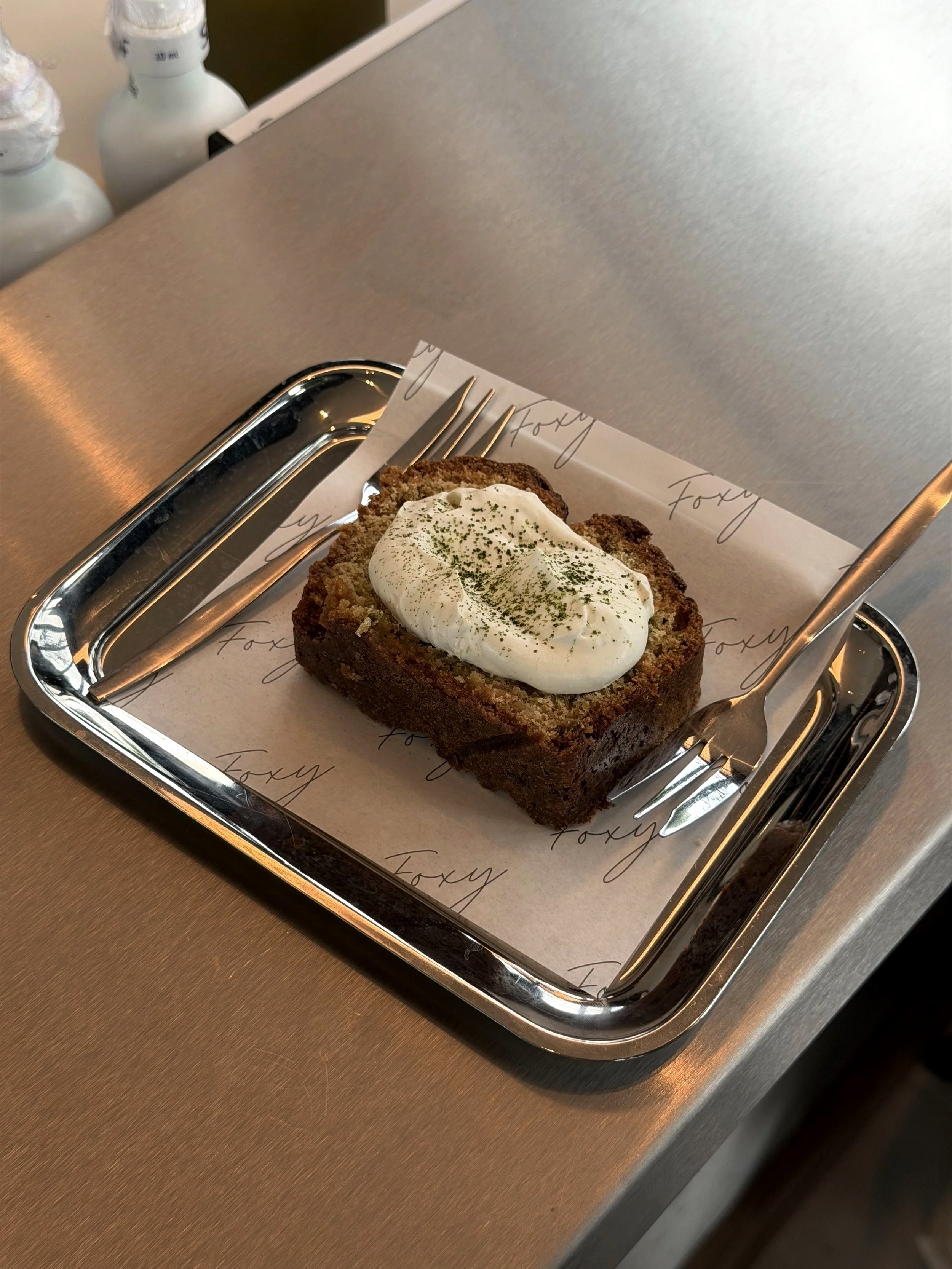 A tray with a slice of loaf cake topped with a dollop of whipped cream, garnished with green powder, on branded parchment paper with cursive writing, on a metal table.