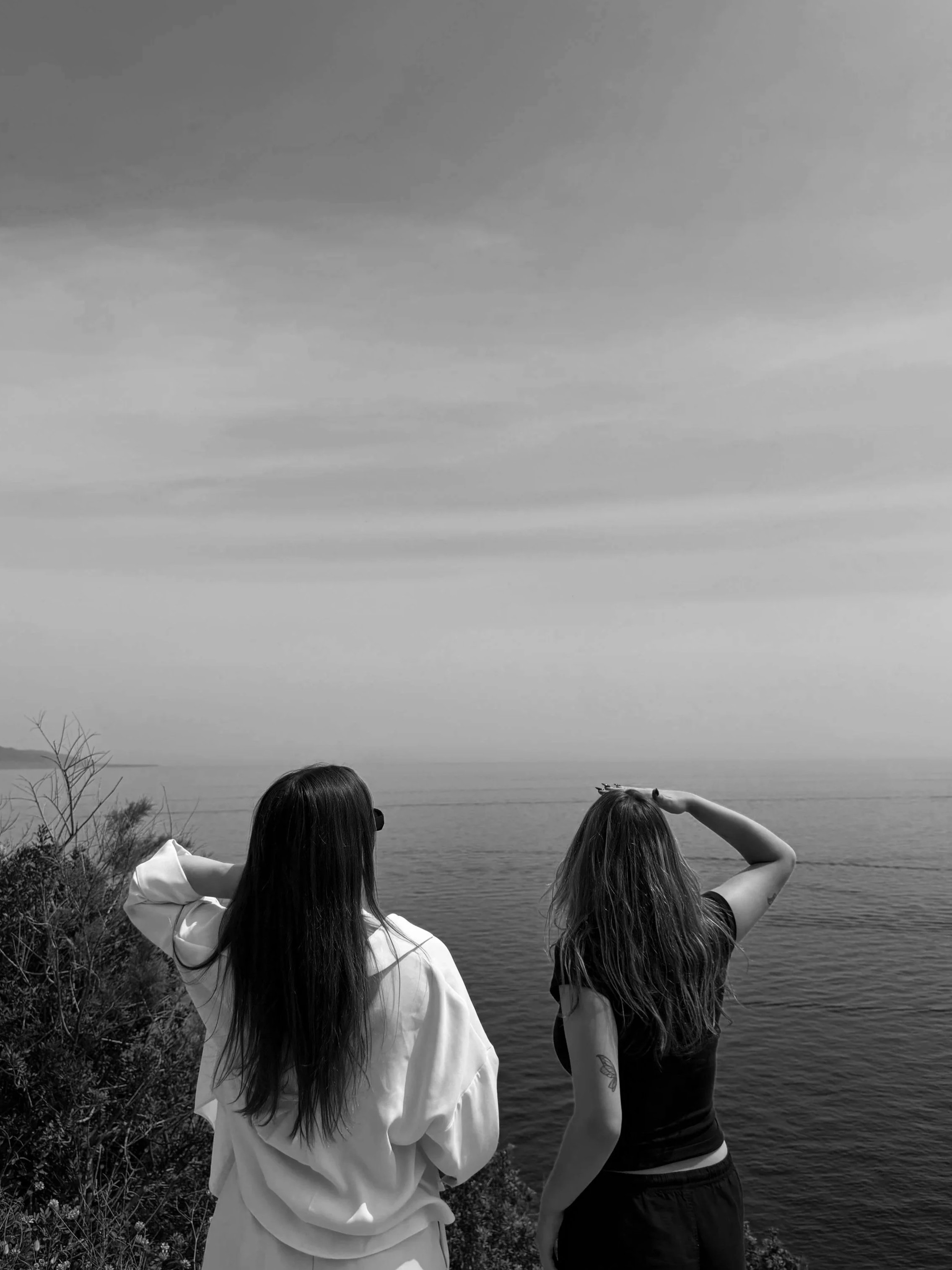 Two women with long hair standing by a body of water, one shielding her eyes from the sun and the other looking out, with hills and trees in the background.