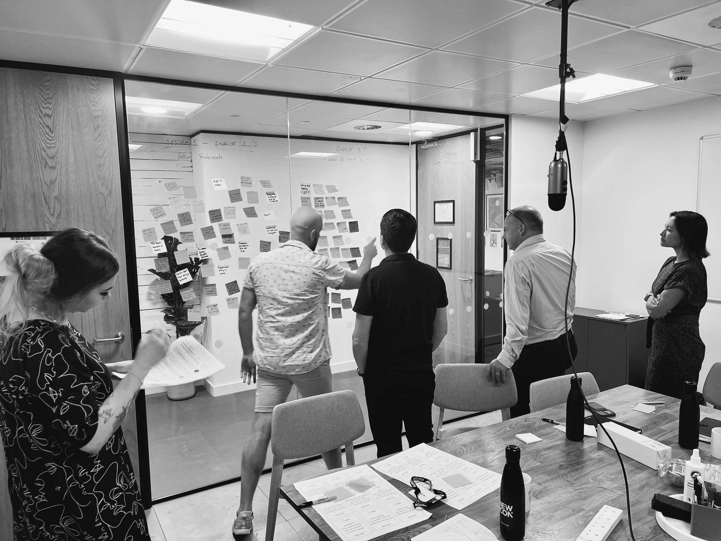 Group of people in a conference room, some standing near a wall with sticky notes, others sitting at a table with papers and bottles, engaging in discussion.