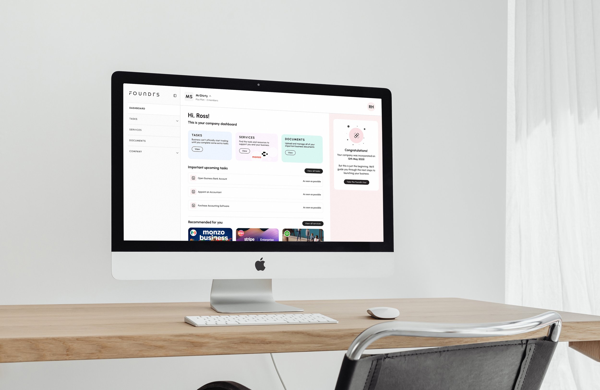An Apple iMac computer on a light wood desk displaying a company dashboard website with menu options on the left and a greeting to Ross on the screen.