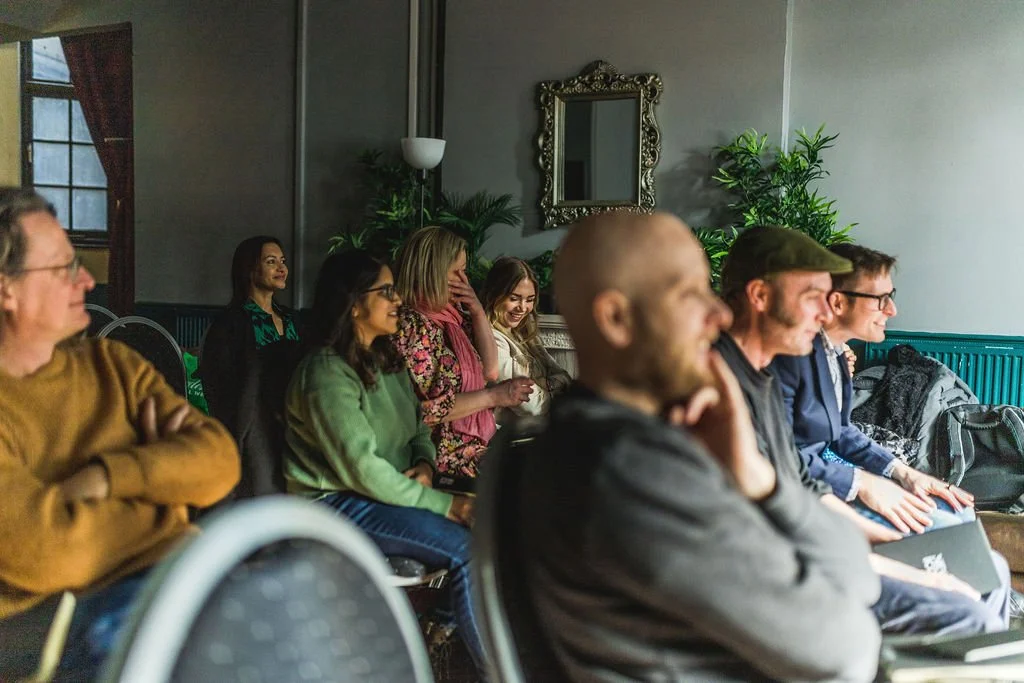 People seated in a conference room, attentively listening to a presentation or speaker, with greenery and a mirror on the wall behind them.