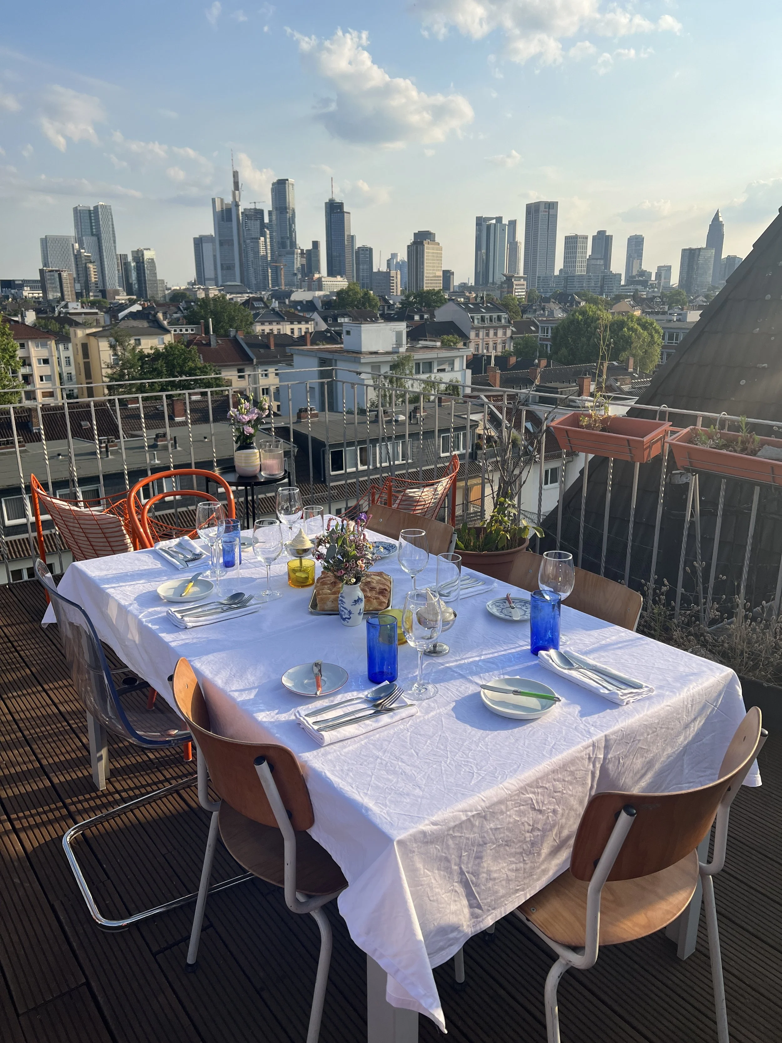 Ein gedeckter Esstisch auf einem Balkon mit Blick auf die Frankfurter Skyline mit Hochhäusern, inklusive Blumen, Gläser, Teller, Besteck und Servietten.