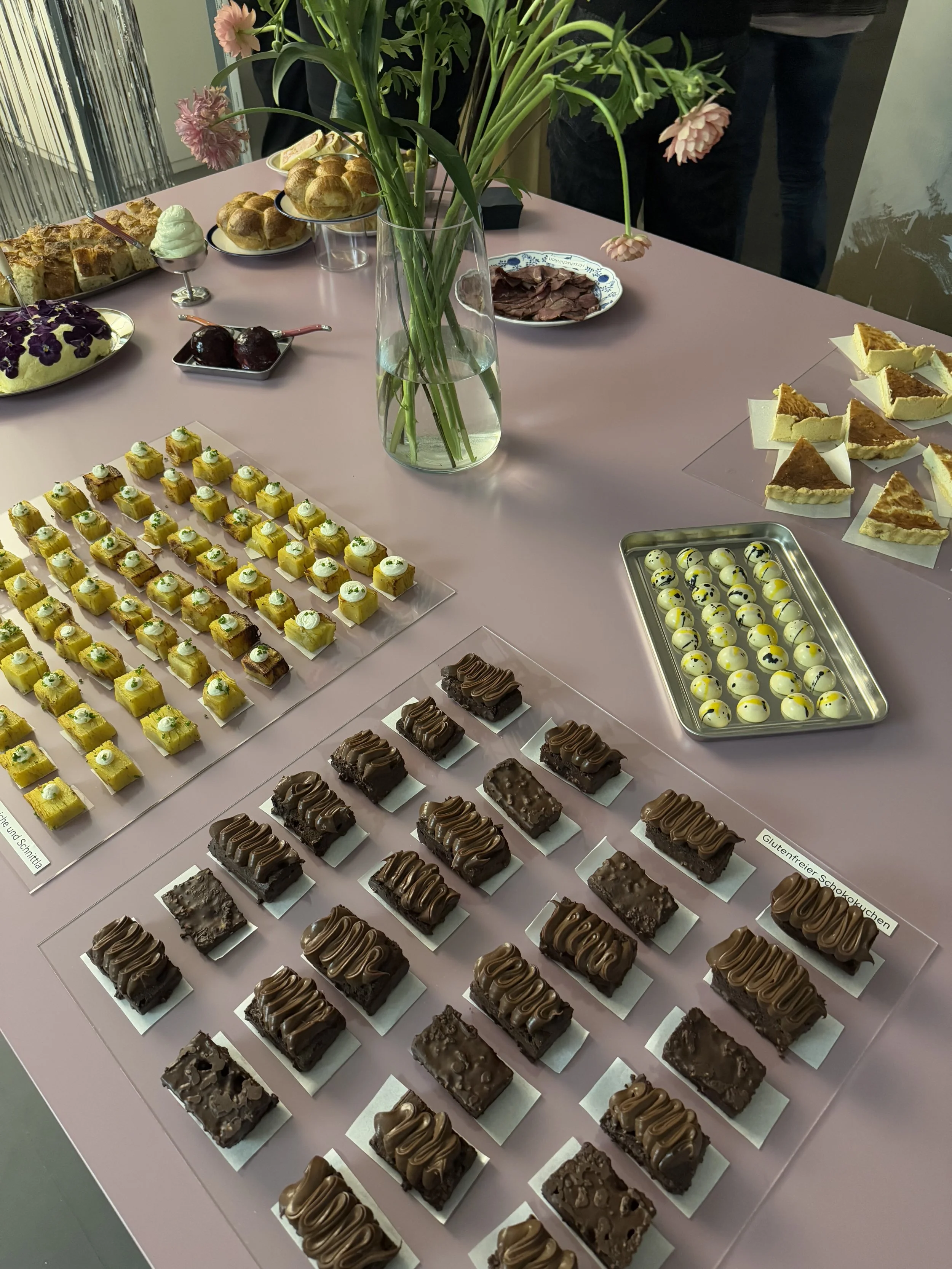 Schokoladenkuchen, Kartoffel-Pavé, Pralinen und ein Blumenstrauß in einer Vase, auf einem pinken Tisch.