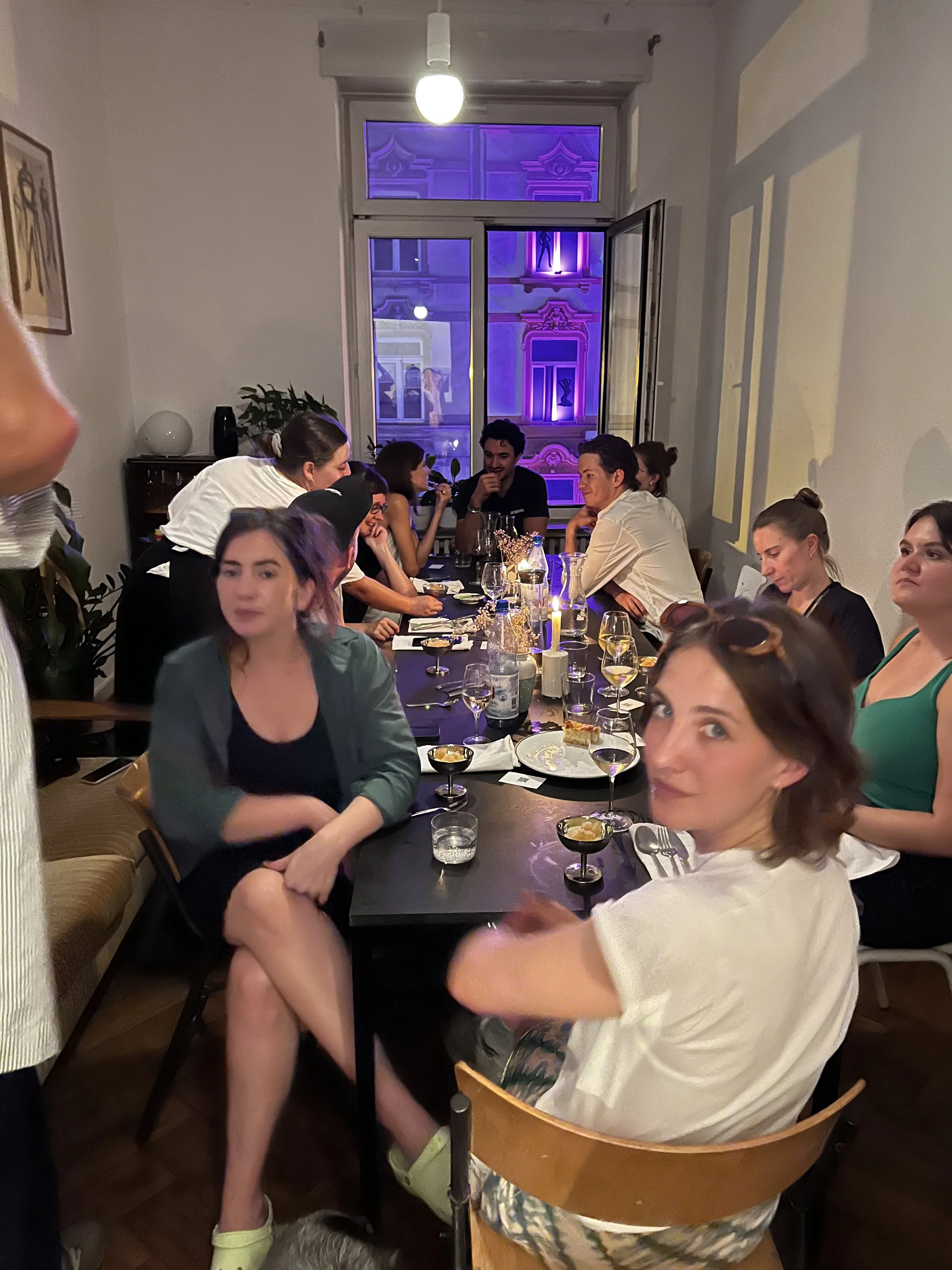Menschen sitzen an einem langen Esstisch bei einem Supper Club, es gibt Wein, Kerzen und Essen, im Hintergrund sind bunte Lichter und die Fassade eines Rotlichtgebäudes durch das Fenster sichtbar.