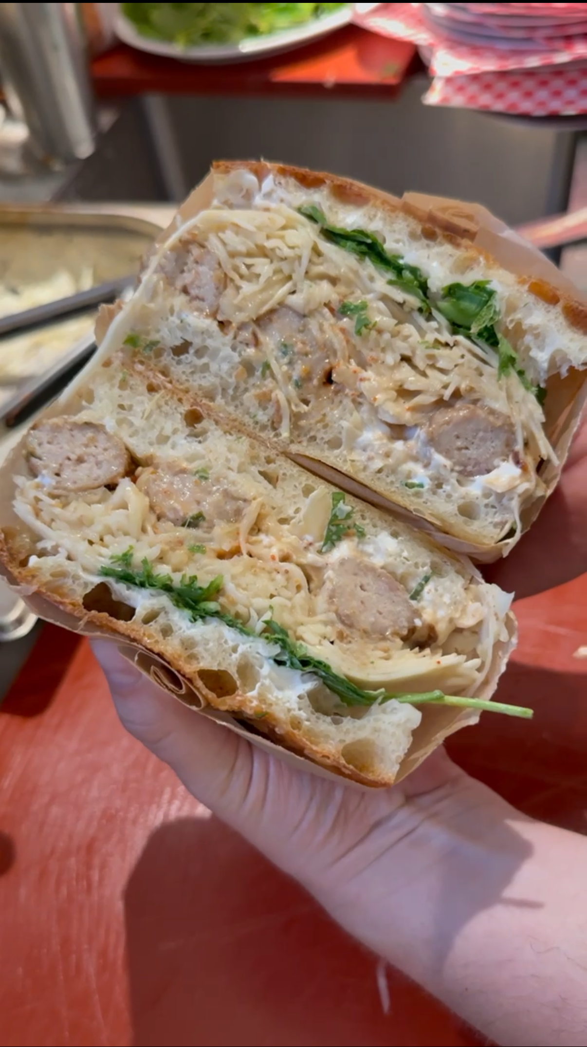 Eine Hand hält ein halbiertes Sandwich mit Tsukune, Krautsalat, frischen Kräutern, auf einem roten Schneidebrett.